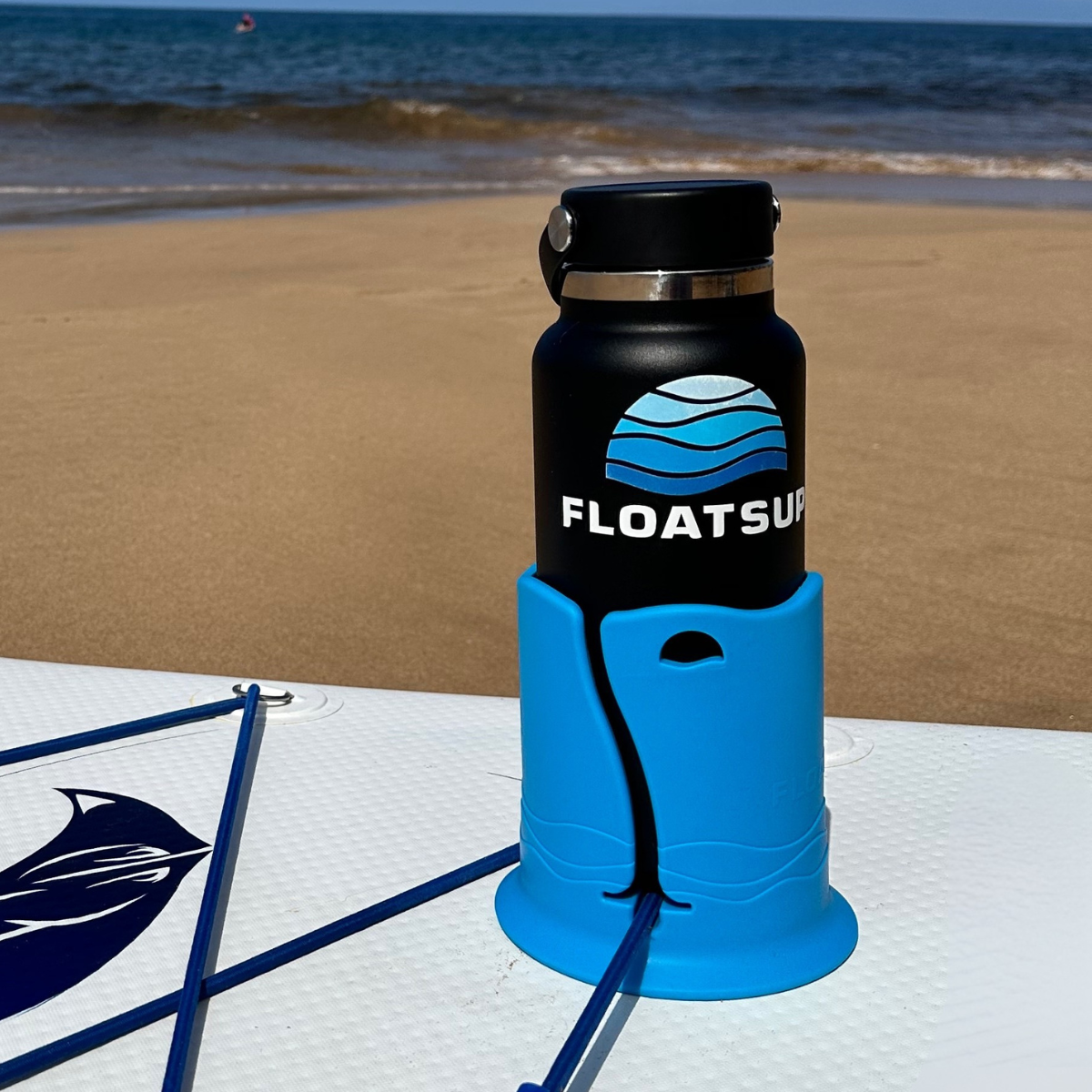 A black water bottle with the blue Floatsup logo on it is in a blue cup holder on a white paddleboard against a beach background.