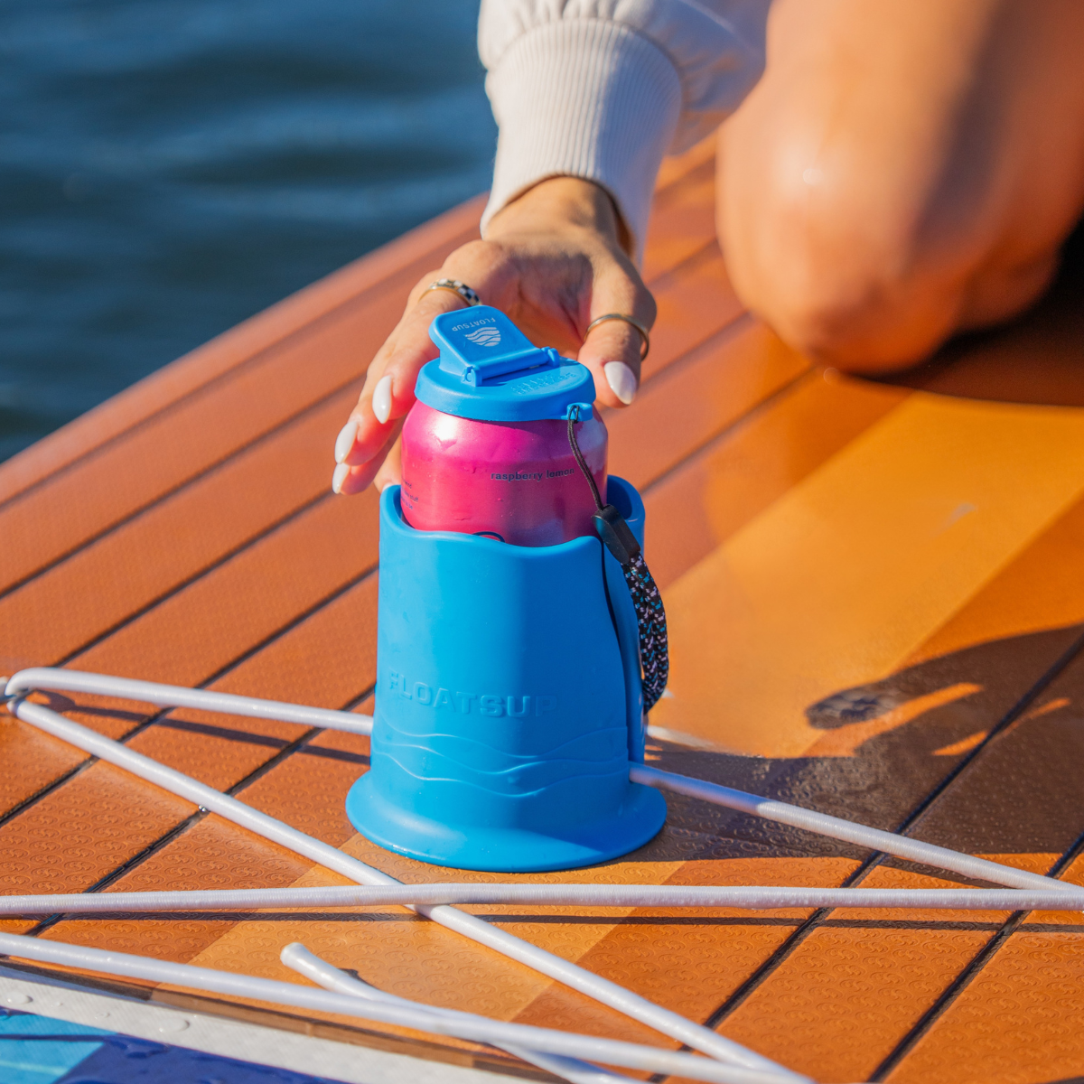 A hand reaching for a pink can in a bright blue cup holder on top of a brown striped paddleboard in water.