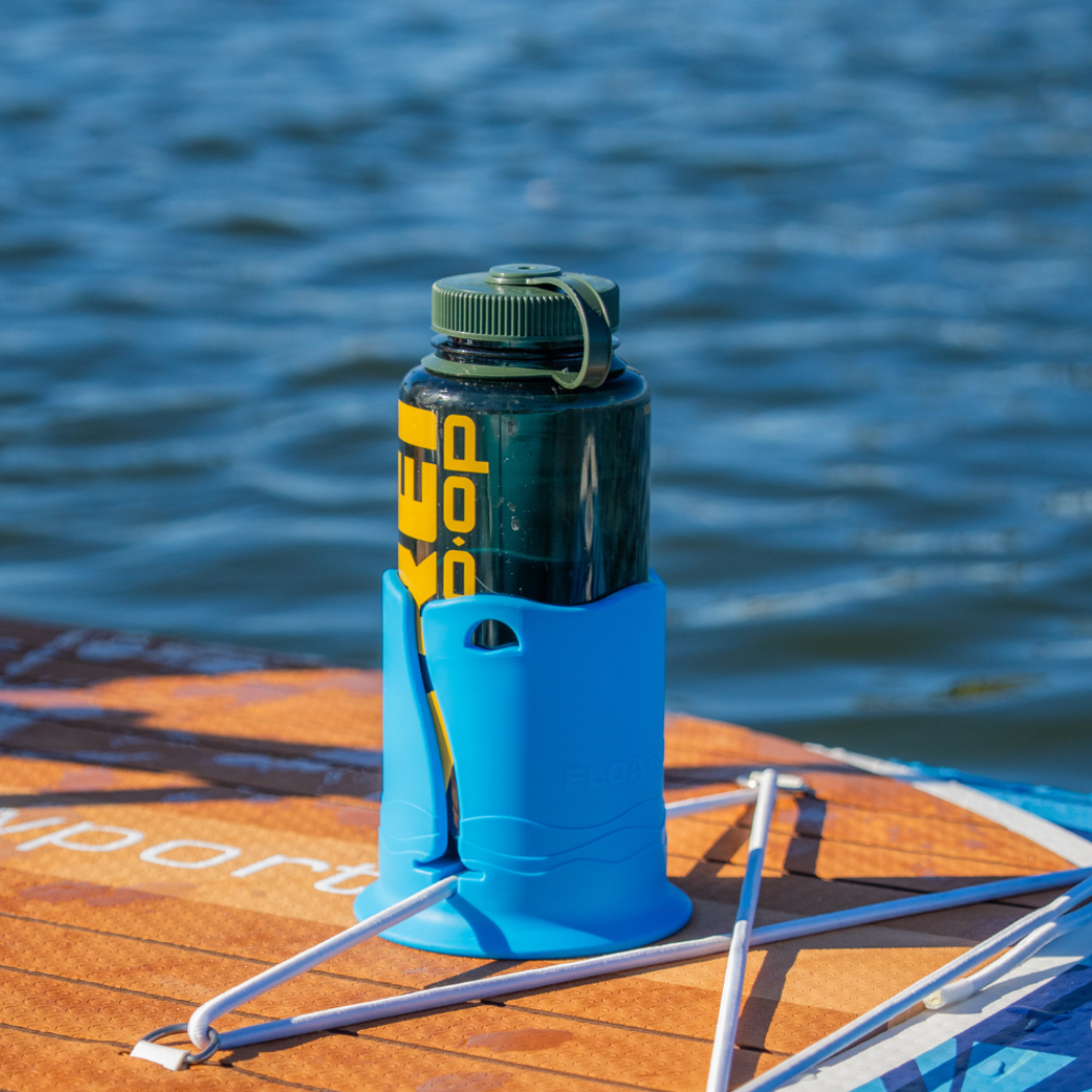 A green translucent water bottle in a blue cup holder attached to the top of a brown paddleboard, with blue water in the background.
