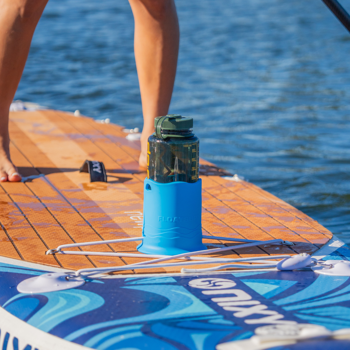 A person is standing on a paddleboard with a green translucent water bottle in a blue cup holder attached to the top in the foreground. The person is paddling with water in the background.