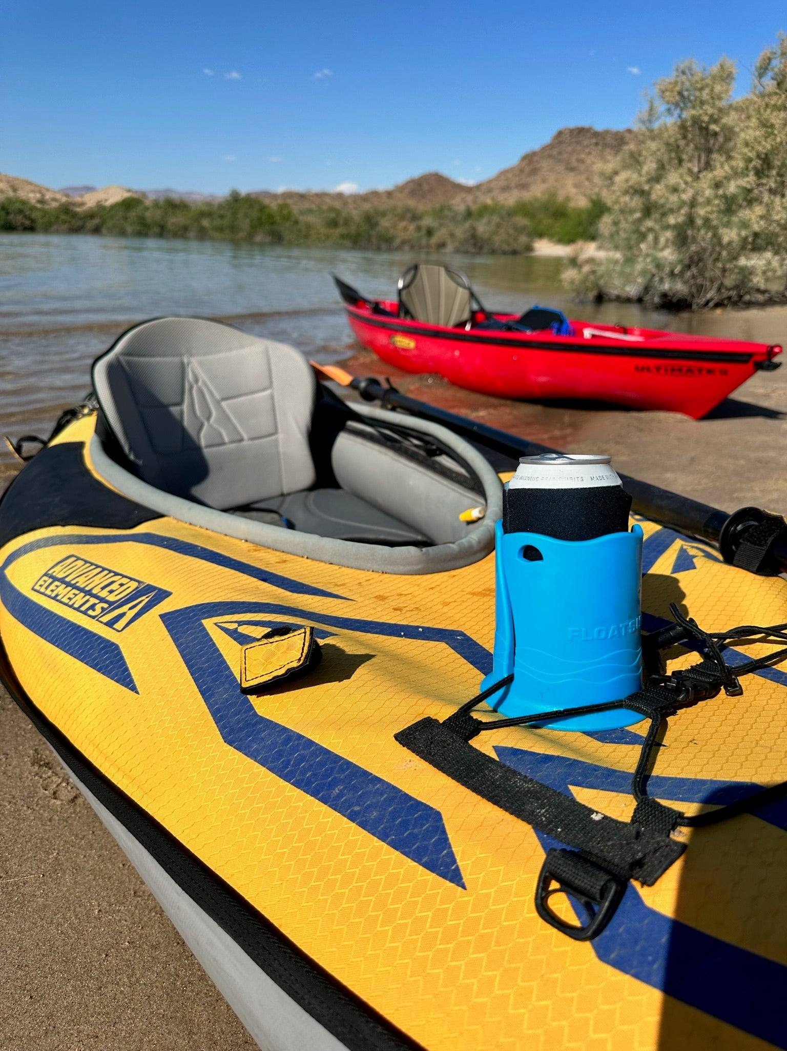 A can in a blue cup holder on top of a yellow inflatable kayak with a gray seat on a sandy shore with a red kayak and mountains in the background.