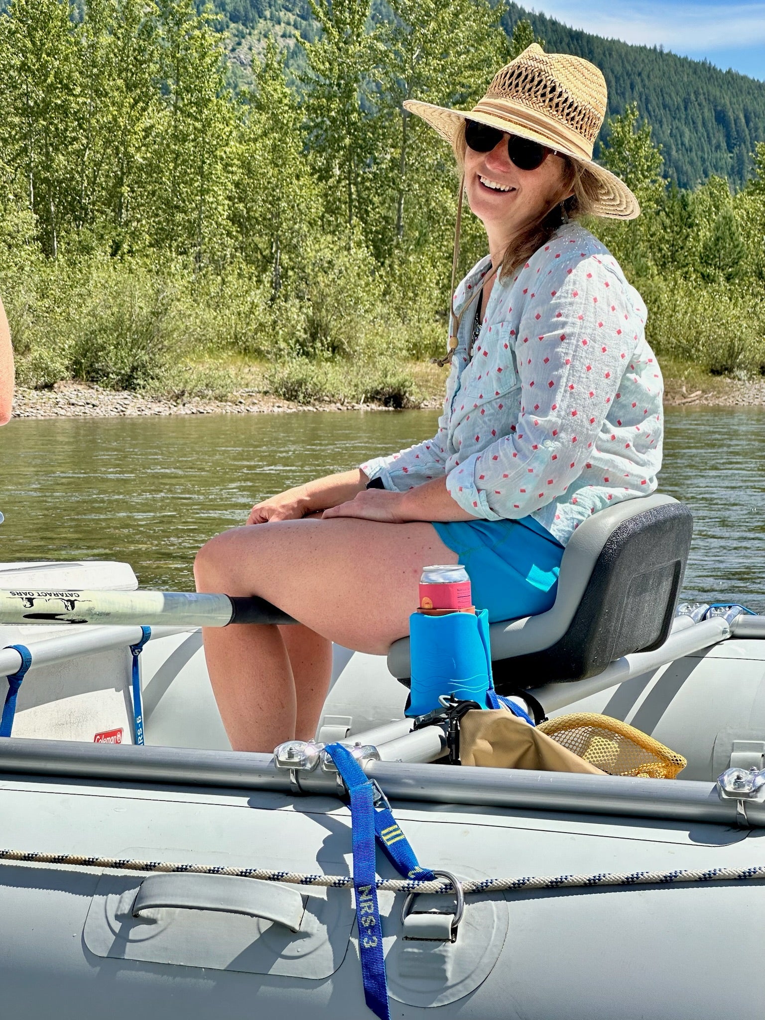Woman sitting in a boat on a river with trees and mountains in the background. She has a blue cup holder with a can inside next to her.