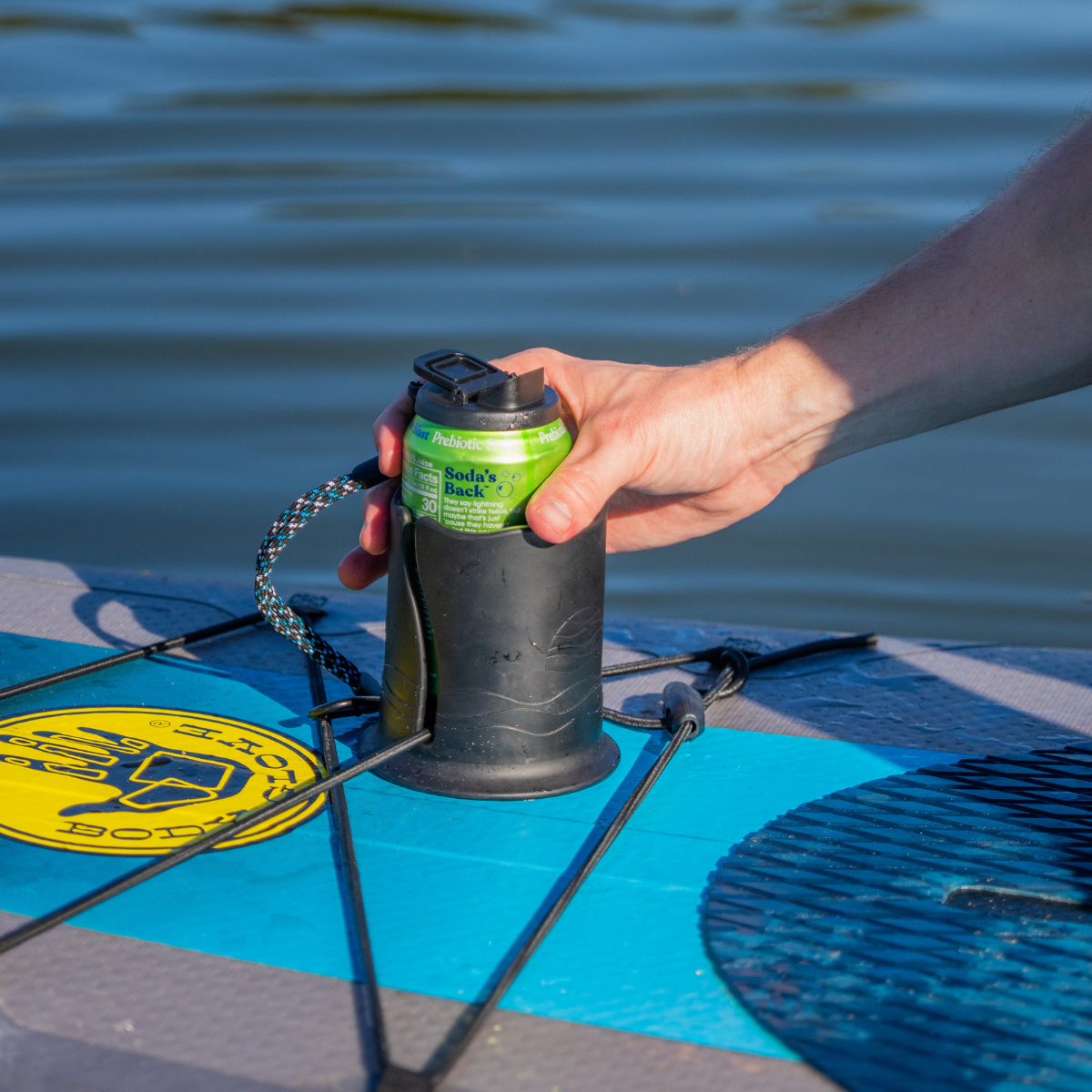 A hand holding a green can inside of a black cup holder on a paddleboard with water in the background.