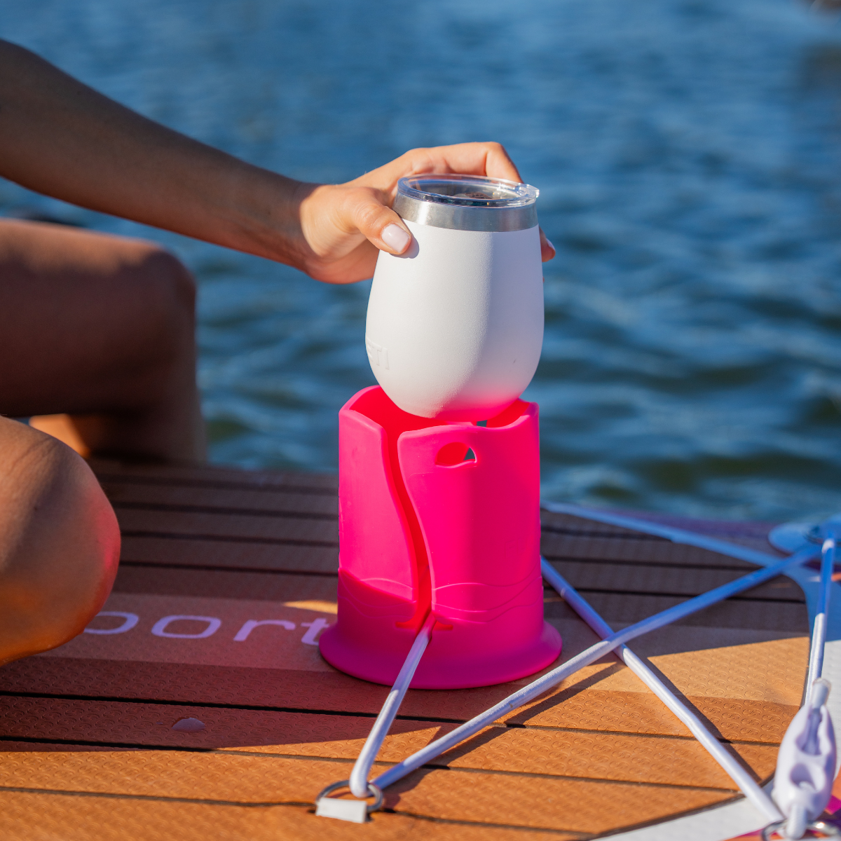 A hand is placing a short white tumblr into a pink cup holder attached to a brown paddleboard on water.