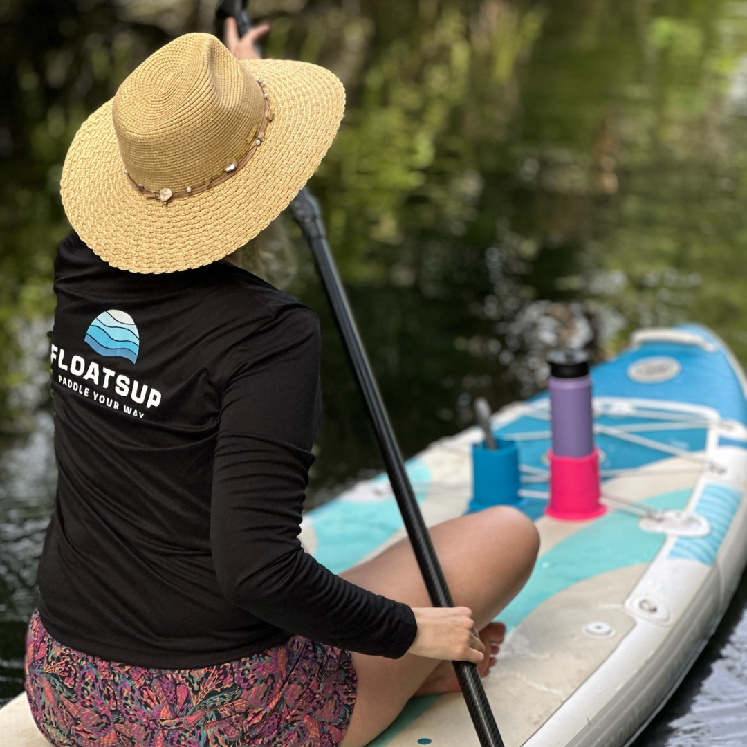 A woman sitting on a paddleboard in a beige hat and long-sleeved black shirt with the Floatsup logo on the back, paddling with blue and pink cup holders in front of her attached to her paddleboard on the water.