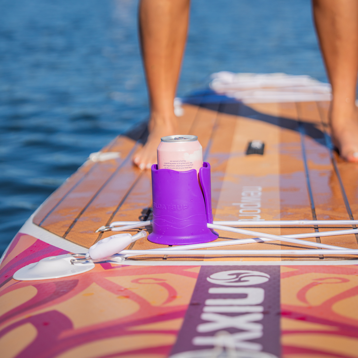 Pink can in a purple cup holder attached to a brown paddleboard, with a person standing on it and water in the background.