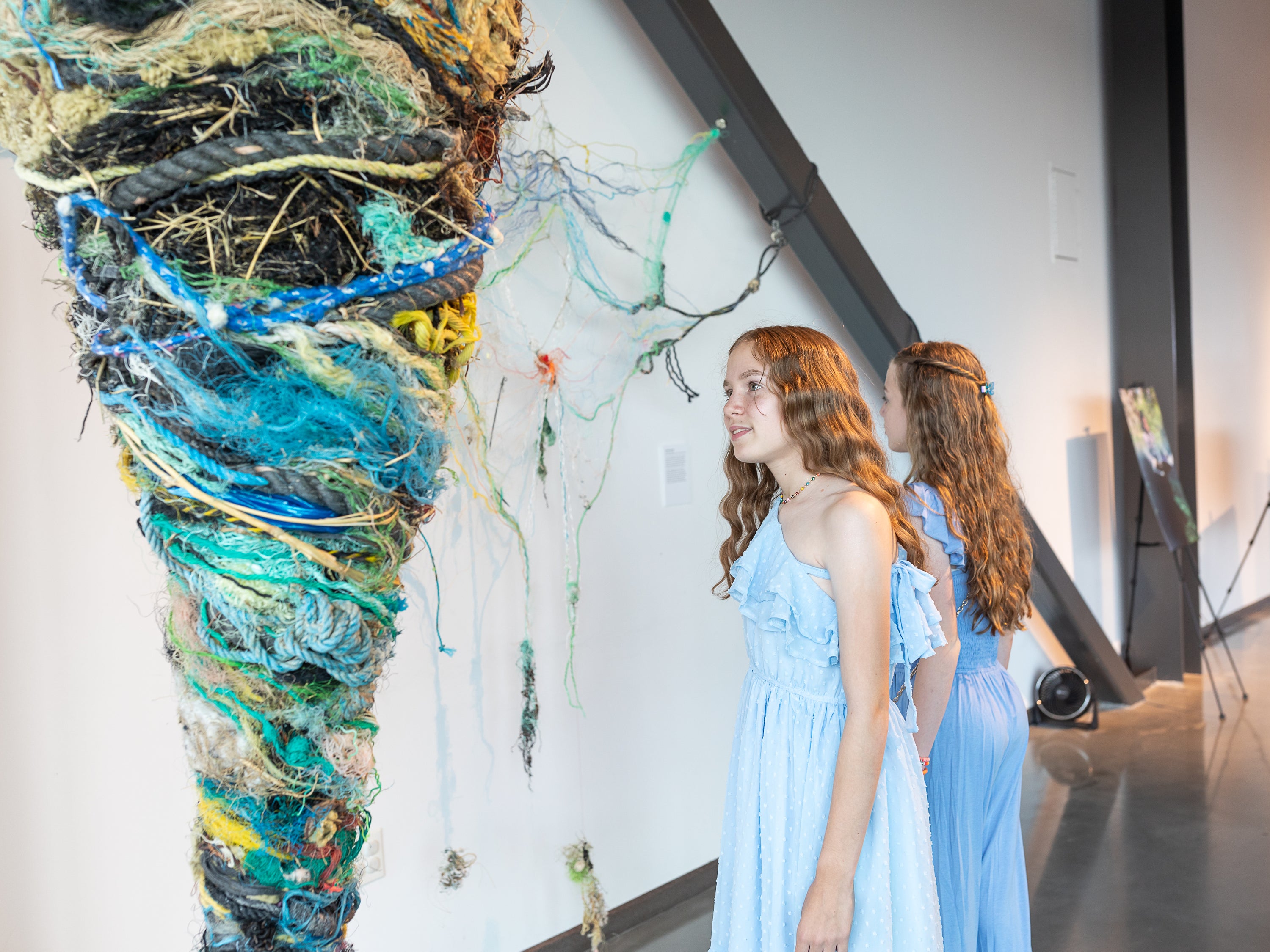Two young girls in light blue dresses with long light brown hair look at an abstract sculpture in an indoor art gallery with white walls. The sculpture is an upside down cone made of various ropes, twine, and nets.