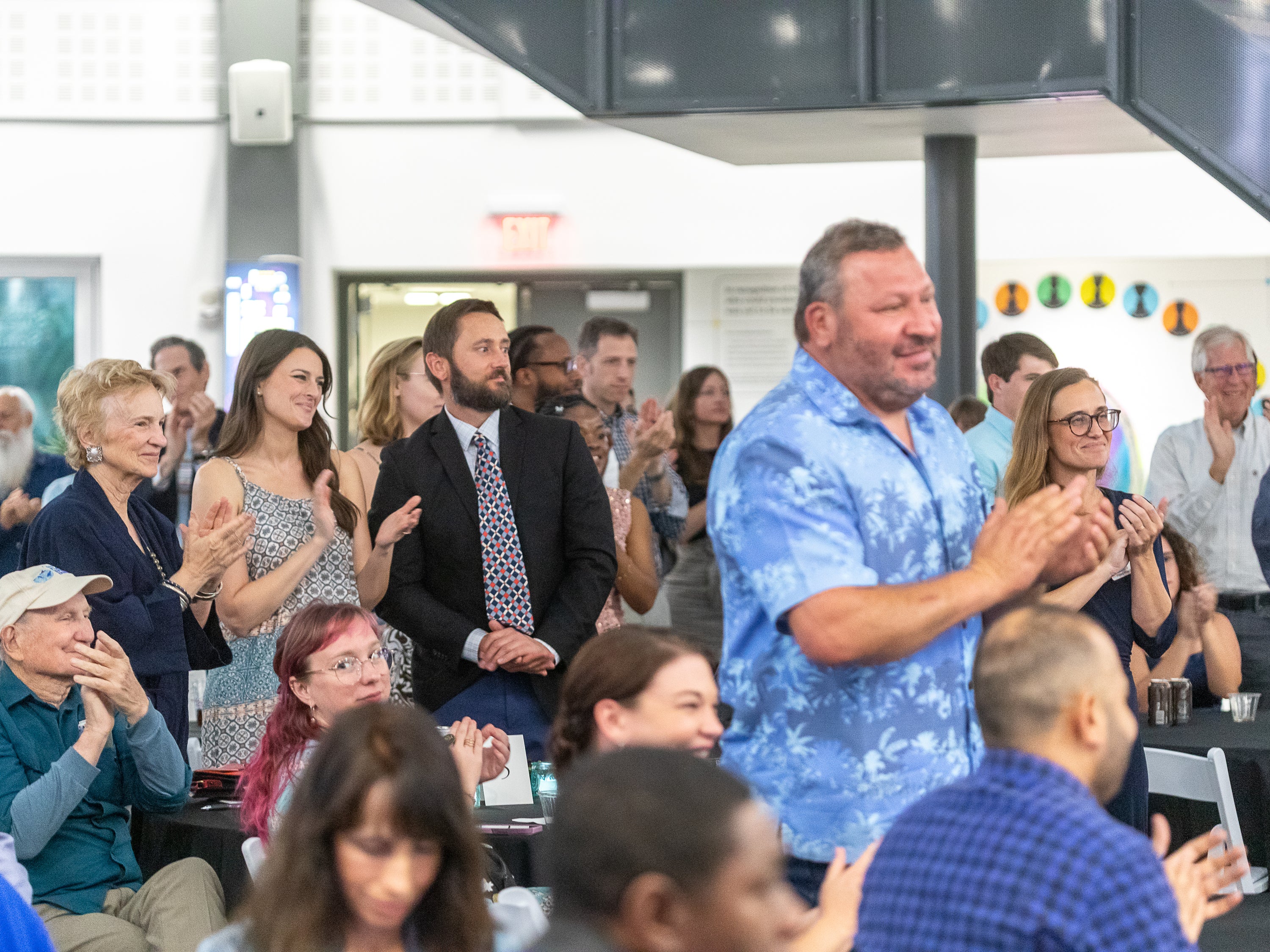 A crowd of people clapping indoors, with a man in a blue patterned short-sleeve shirt standing prominently in the foreground.