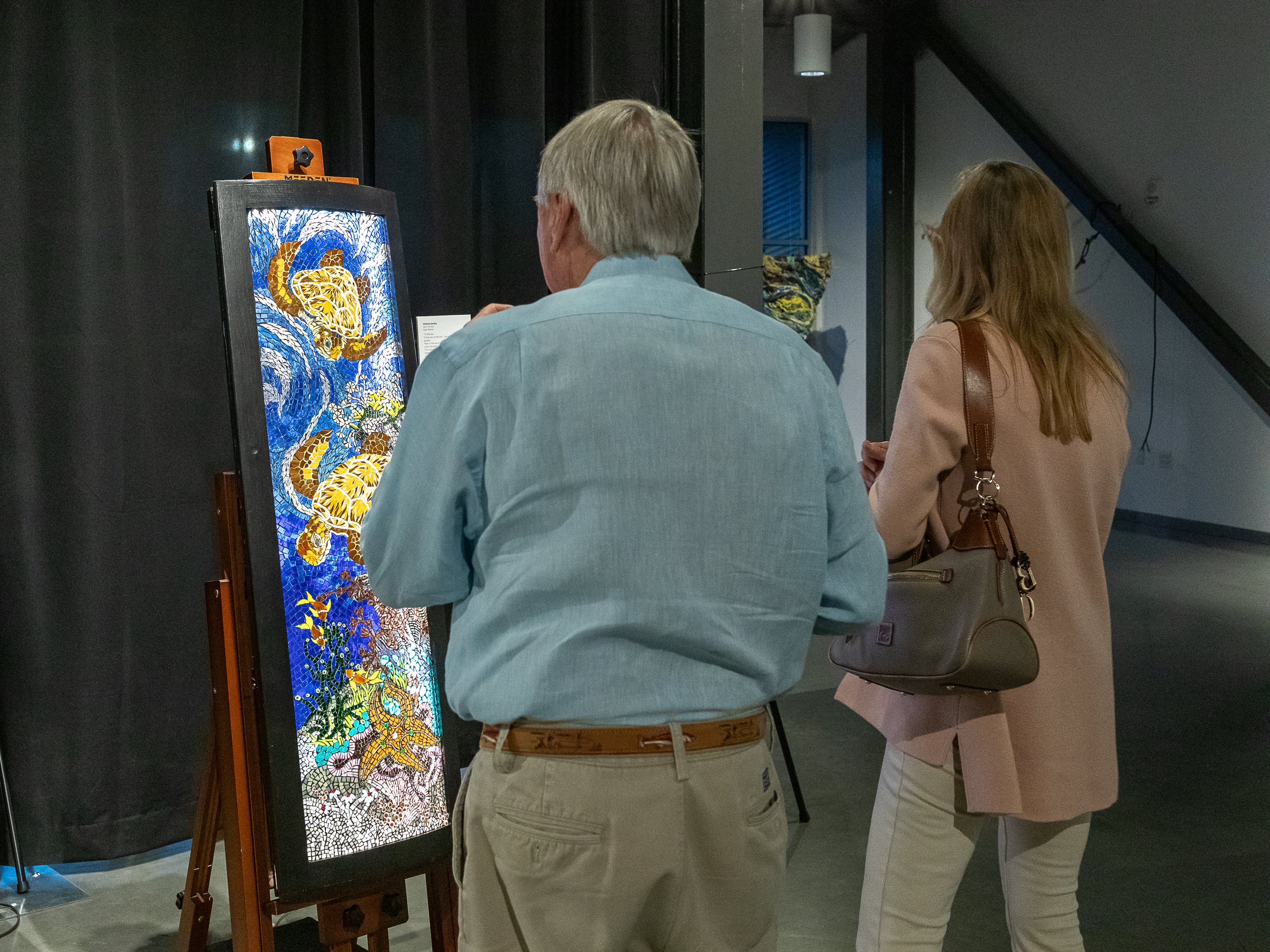 An illuminated mosaic art piece depicting sea turtles is displayed on an easel in an indoor art gallery, with an older man and a woman viewing it from behind.