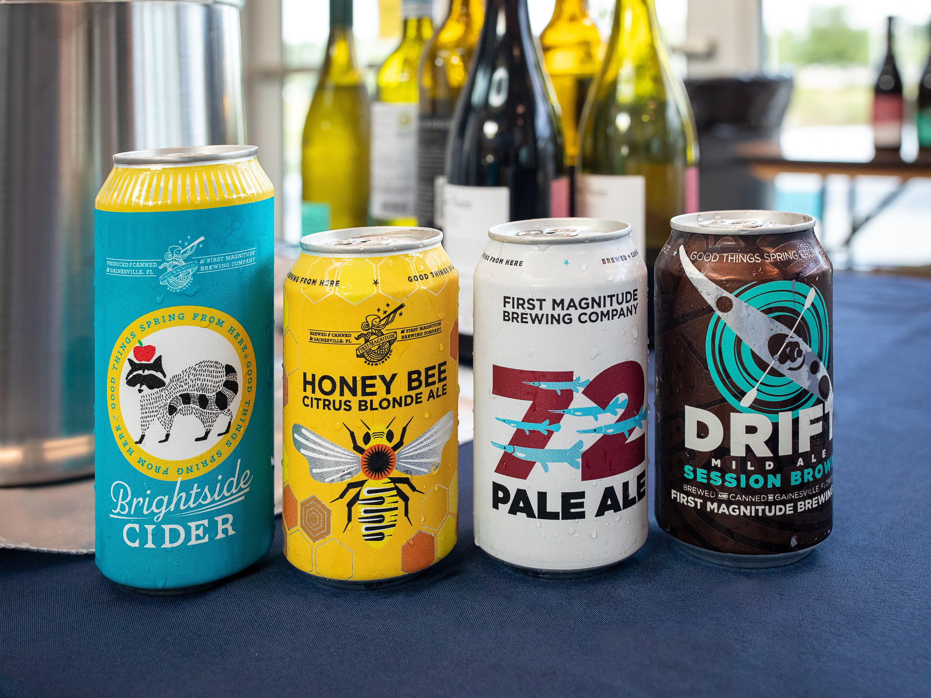Four craft beer cans lined up on a dark blue tablecloth in front of wine bottles. The cans are labeled Brightside Cider, Honey Bee Citrus Blonde Ale, 72 Pale Ale, and Drift Mild Ale, with First Magnitude Brewing Company visible on all the labels.