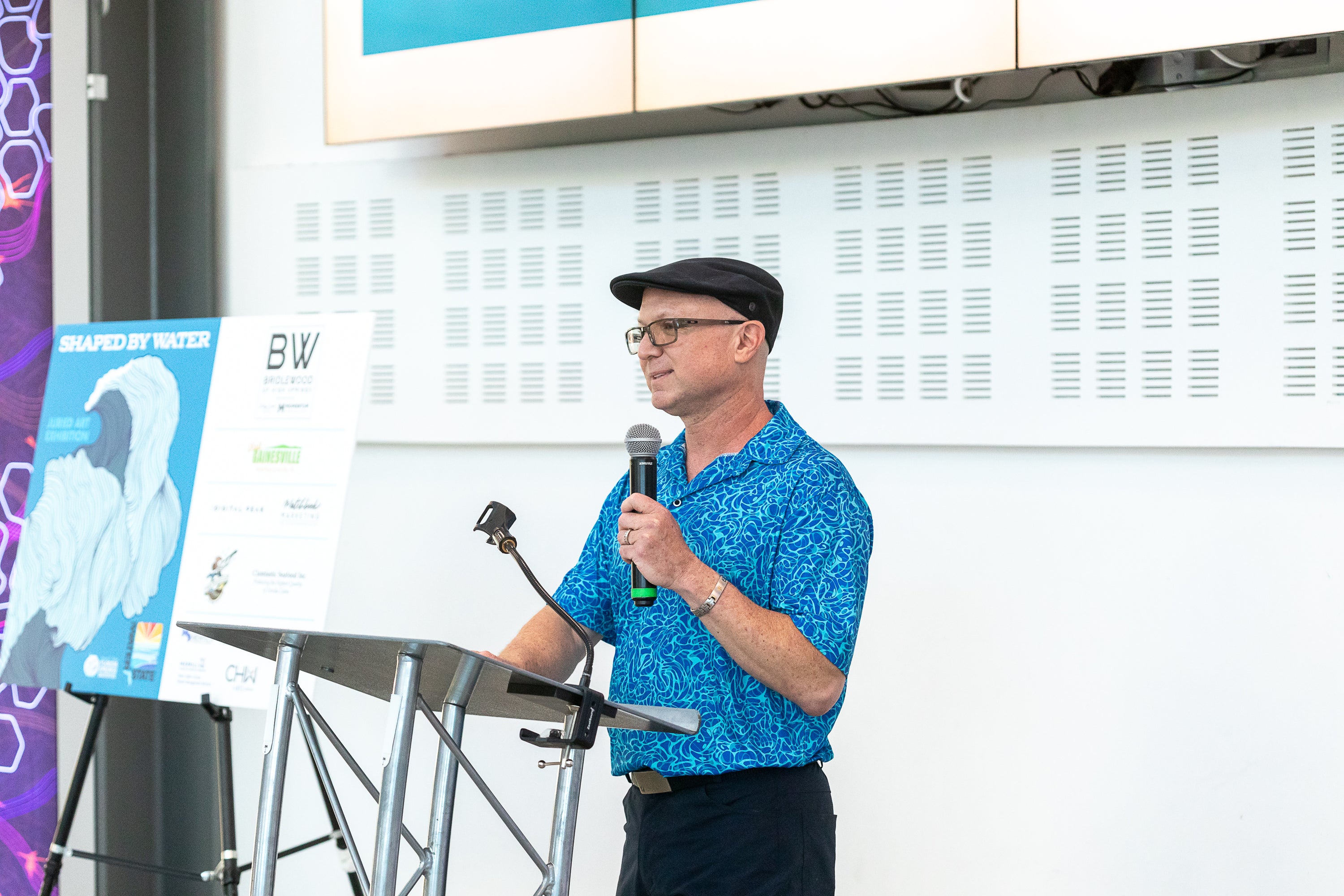A man wearing a blue patterned short-sleeve shirt, black cap, and glasses stands at a podium holding a microphone during a presentation. A branded presentation board is visible to his left against a white wall, that says 'Shaped By Water'.