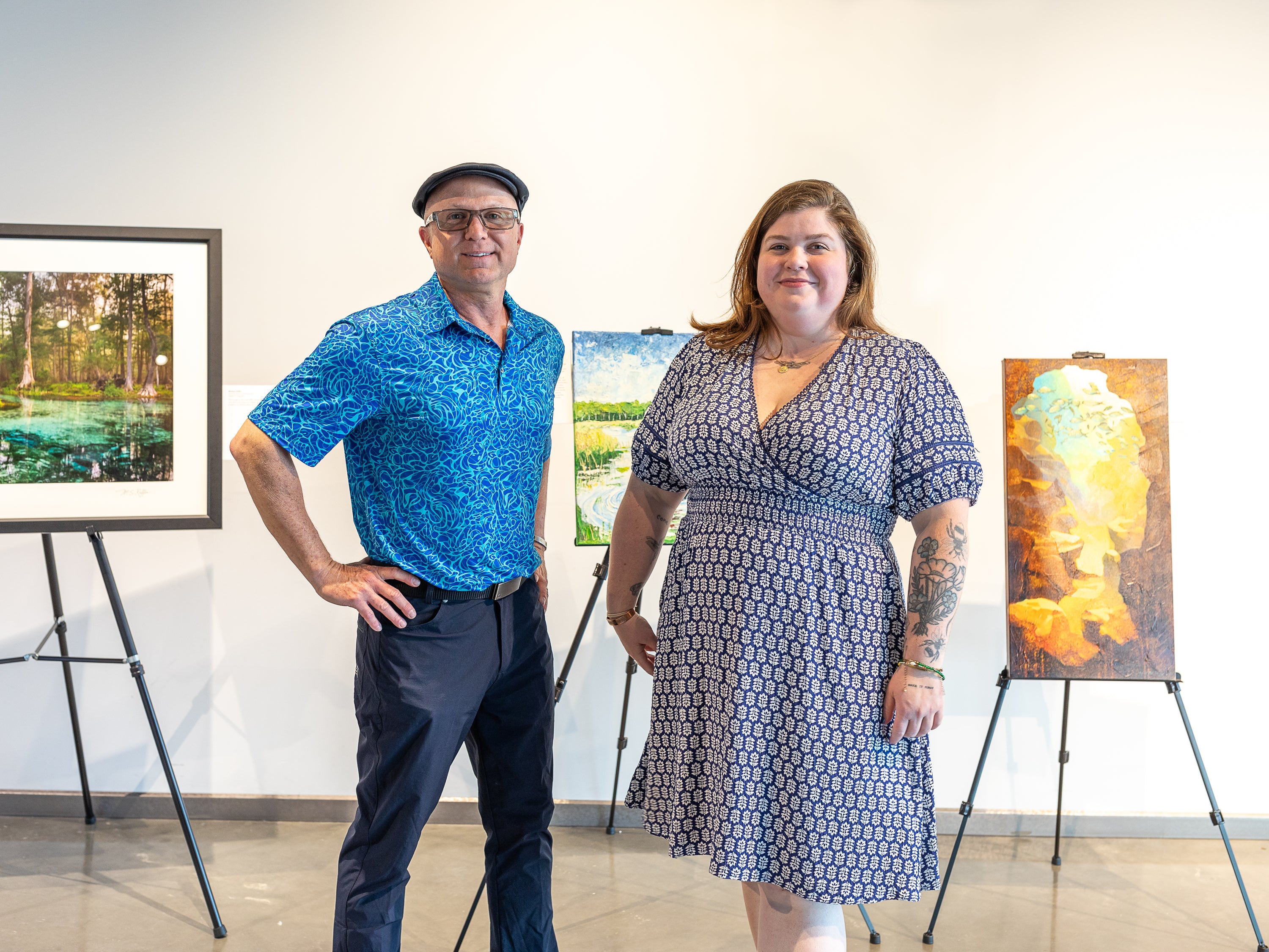 A man and a woman standing in an art gallery space with framed and canvas artworks on easels behind them. The man wears a bright blue patterned short-sleeve shirt, dark pants, and a black cap. The woman wears a navy and white patterned dress.