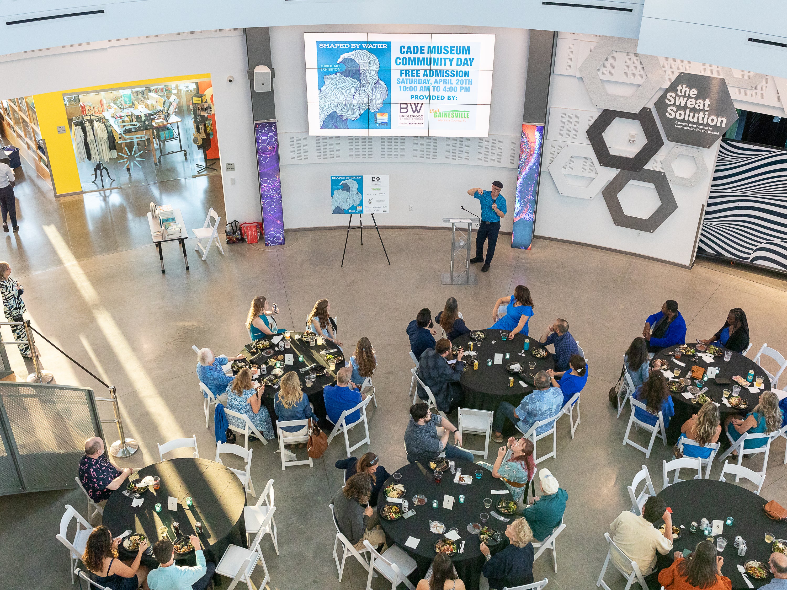 A high-angle view of a community event at the Cade Museum, showing a presenter at a podium and a group of people seated at round tables with food and drinks. A large screen displays 'Cade Museum Community Day'.