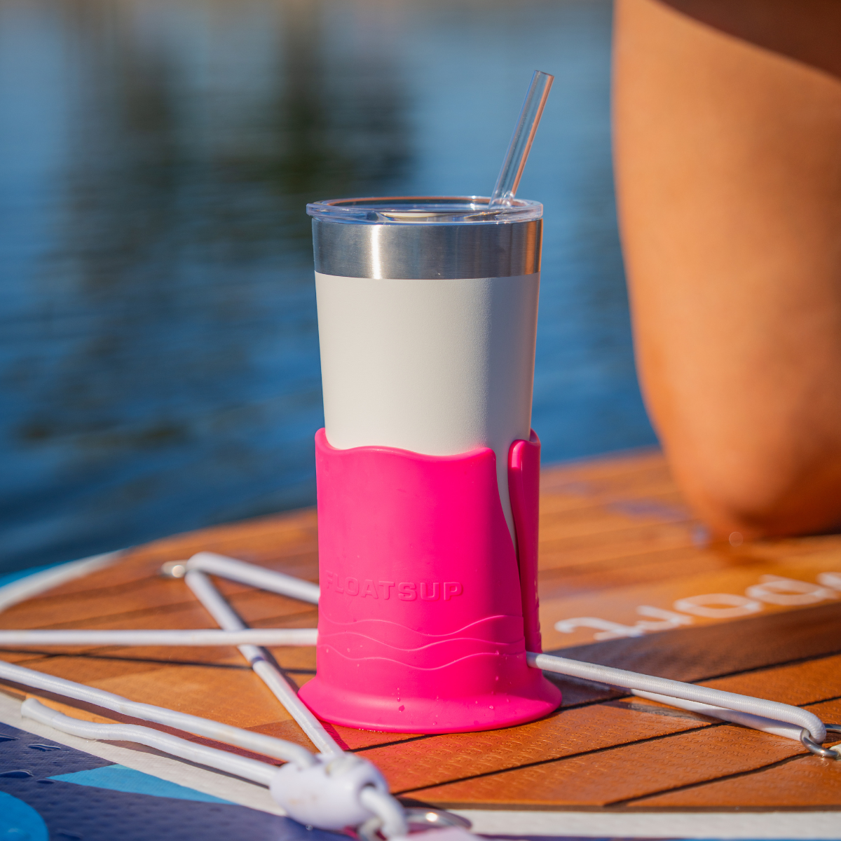 White tumbler with a clear straw in a pink cup holder on top of a brown paddleboard with water in the background.