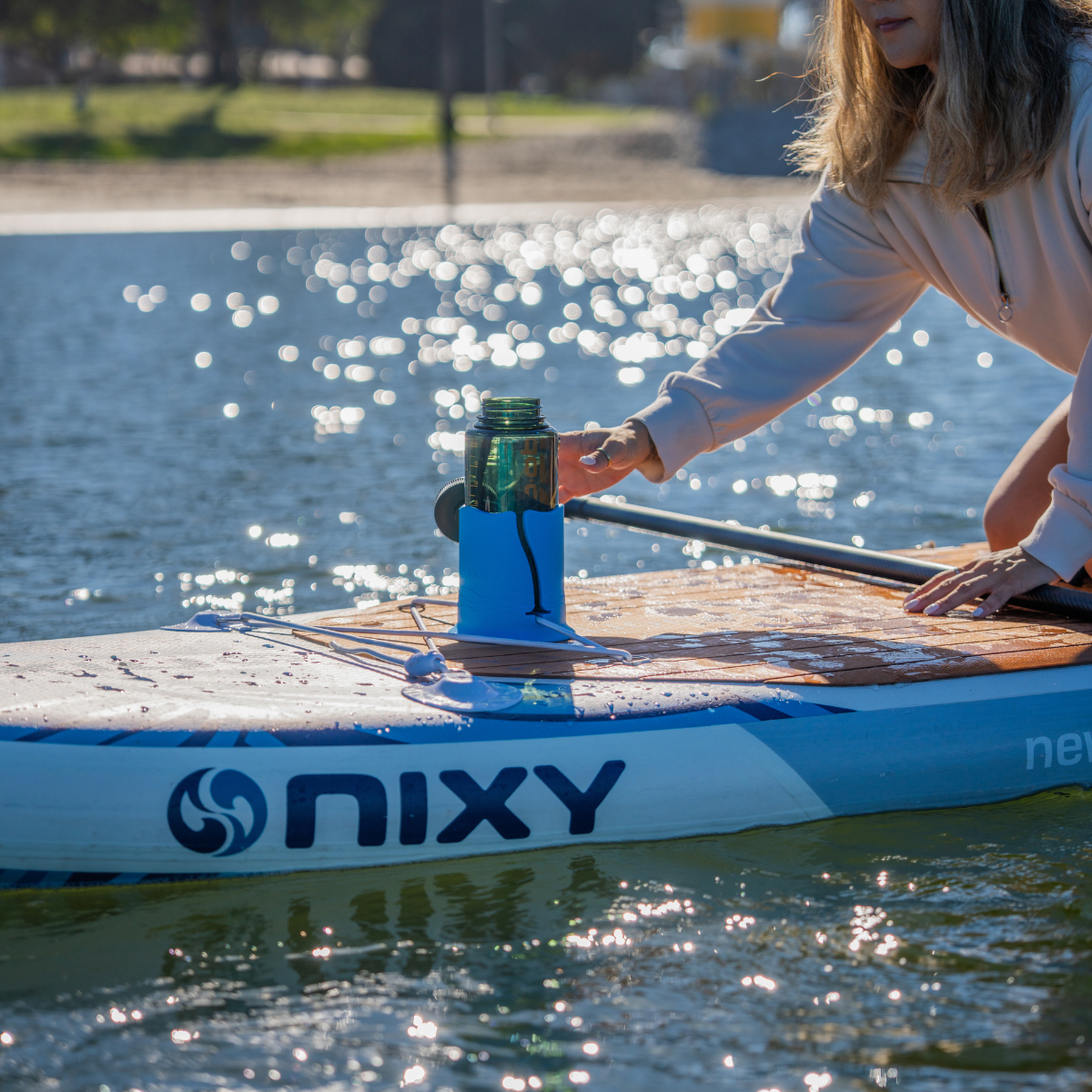 A woman is on a Nixy paddleboard reaching for a green translucent water bottle in a blue cup holder on top of the paddleboard,, with water and grass in the background.