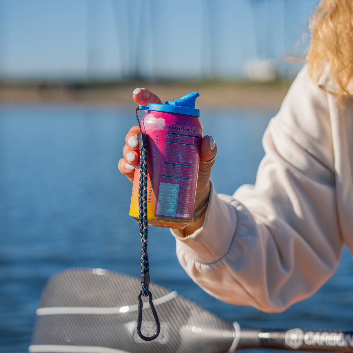A woman's arm holding a pink can with a blue lid in front of water. The lid has a wrist strap and a closable drinking spout.
