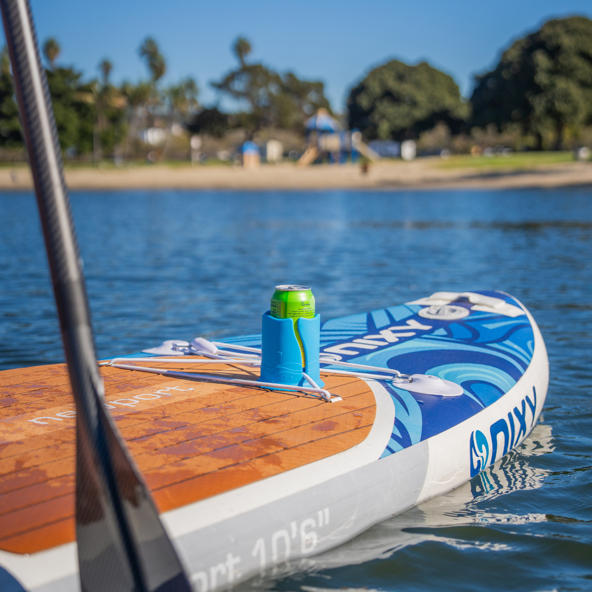 Green can in a blue cup holder on a brown and blue paddleboard in the water, with trees and a blue sky in the background.