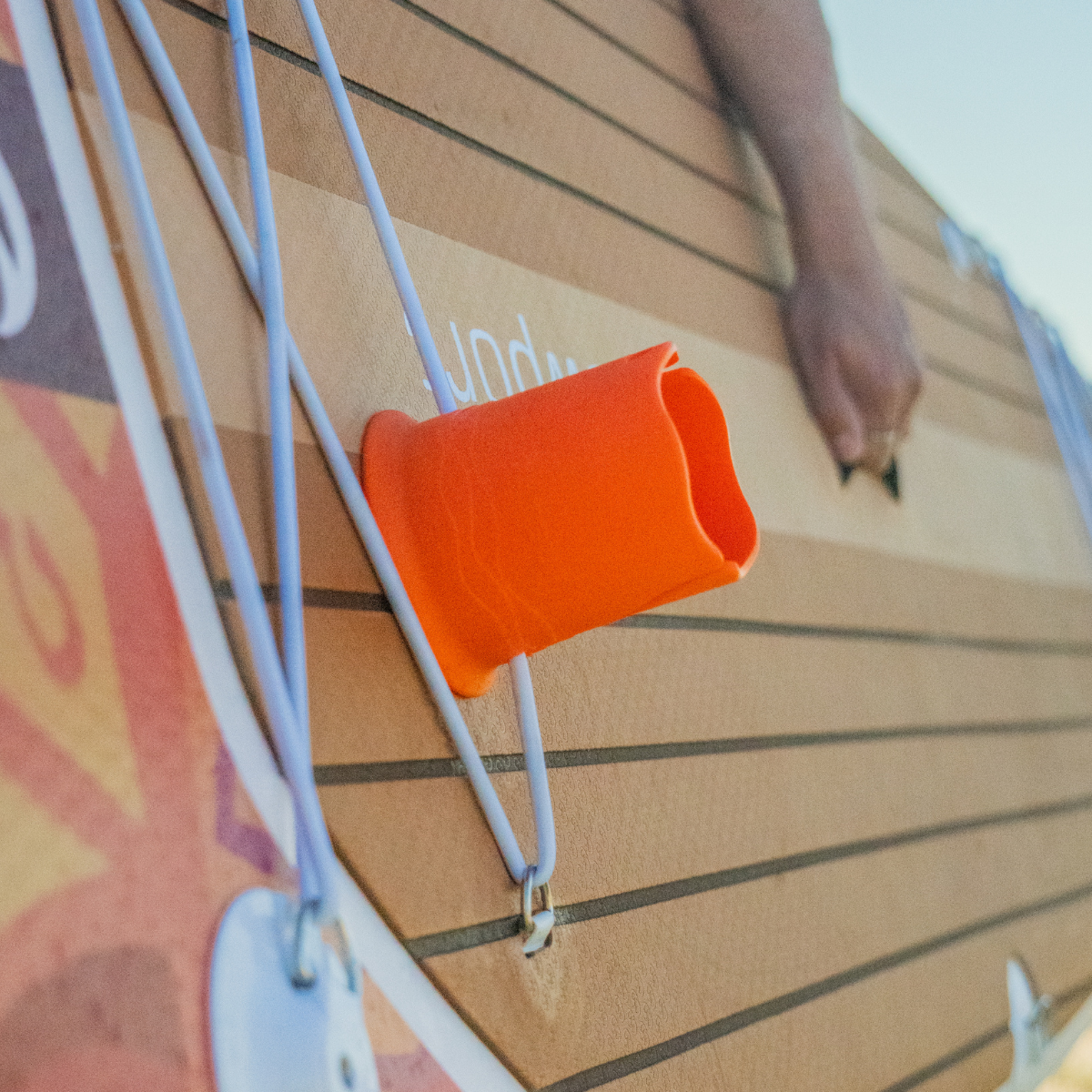 Close-up of a person carrying a brown paddleboard, with a sideways orange cup holder attached by a white bungee cord.