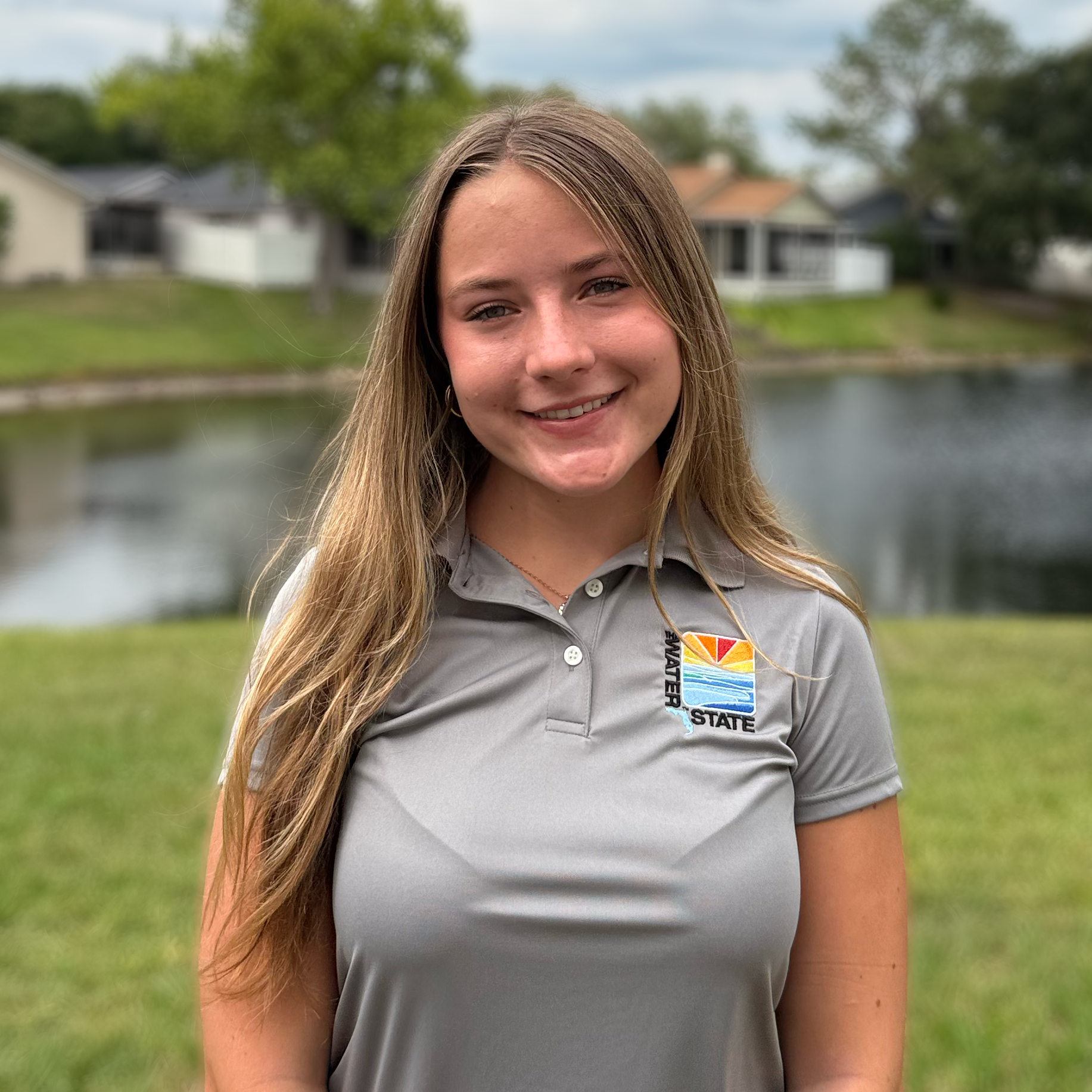 A smiling young woman (Addison) with long light brown hair stands outdoors in front of a pond, wearing a gray polo shirt with a 'Water State' logo on the left chest.