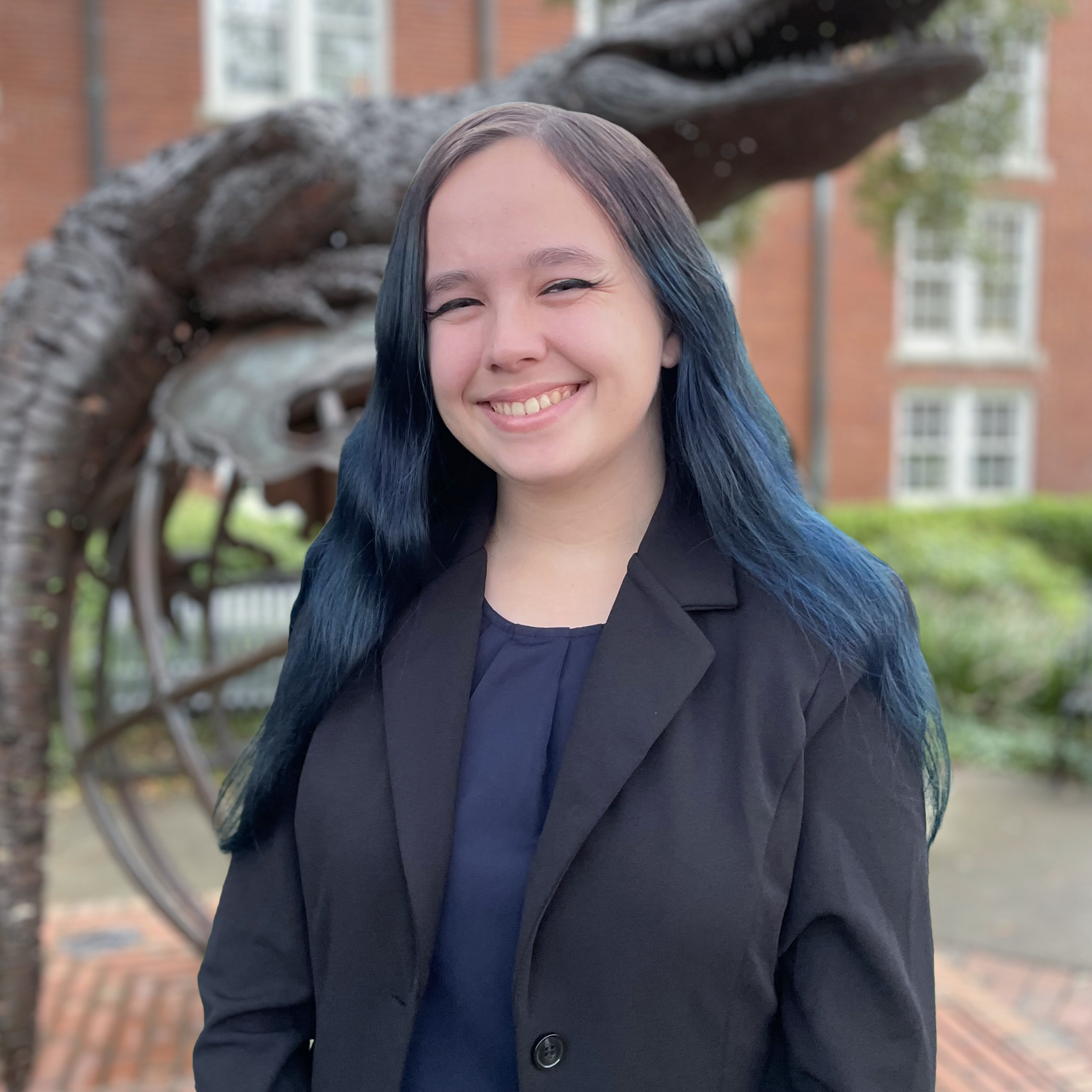 A smiling young woman (Arwen) with long dark blue hair, wearing a black blazer over a navy top, standing outdoors in front of a brick building and a gator sculpture on UF campus.