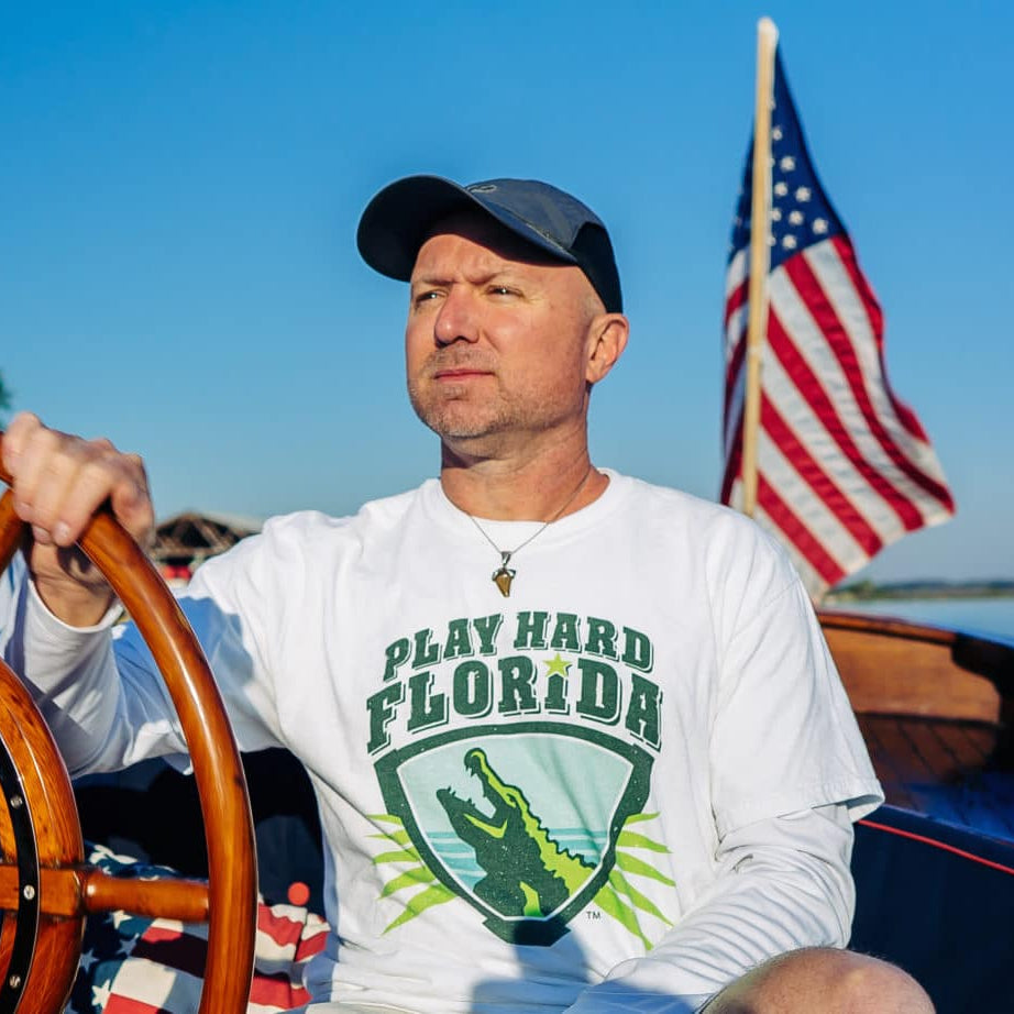 A man (CJ) on a boat wearing a 'Play Hard Florida' shirt with an American flag in the background.