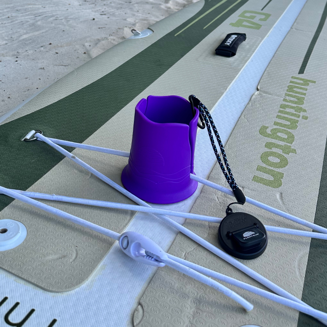 A purple cup holder attached to a green and beige paddleboard on beach sand.