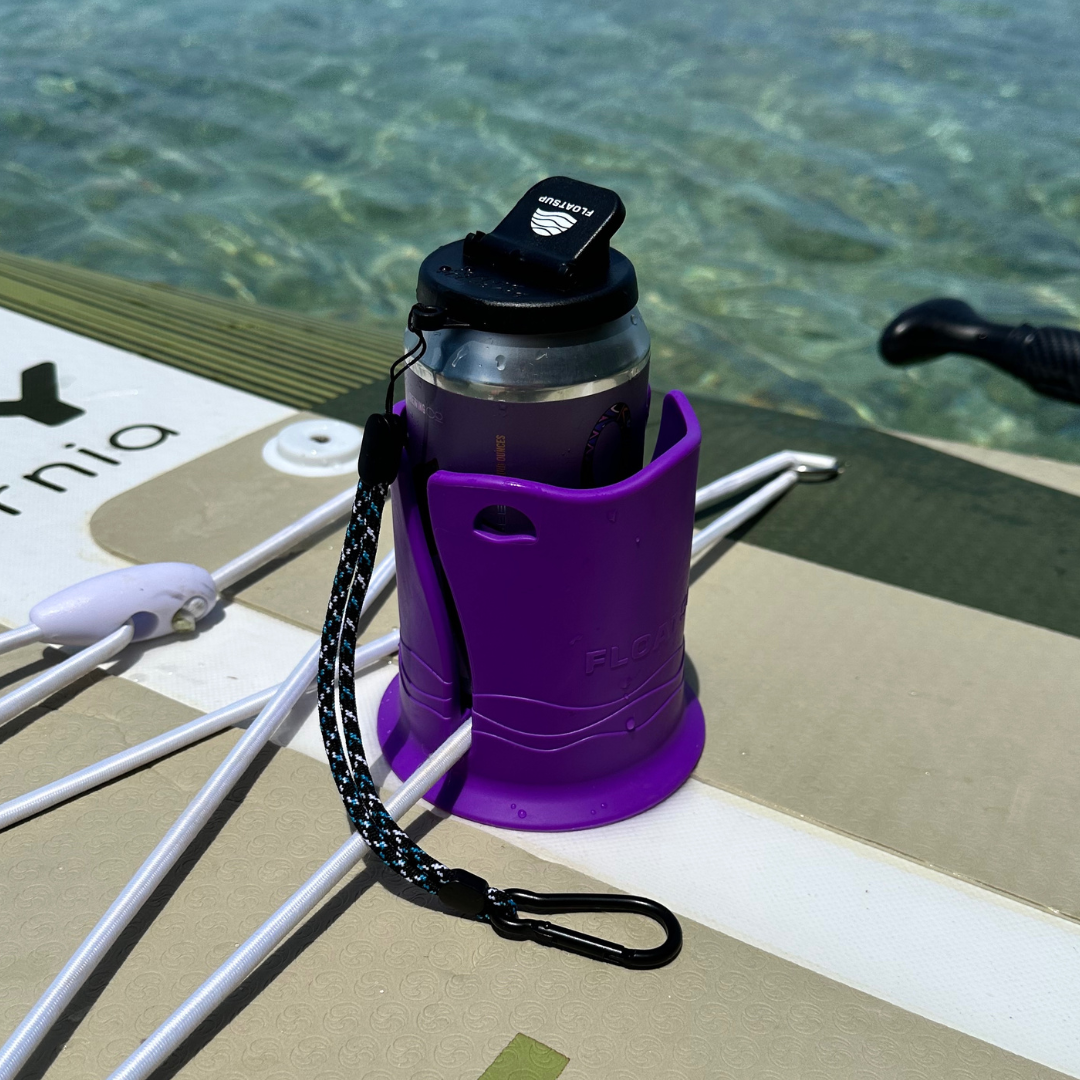 A gray can with a black lid in a purple cup holder on top of a green and beige paddleboard in water. The lid has a wrist strap and a closable drinking spout, and it says 'Floatsup' on top