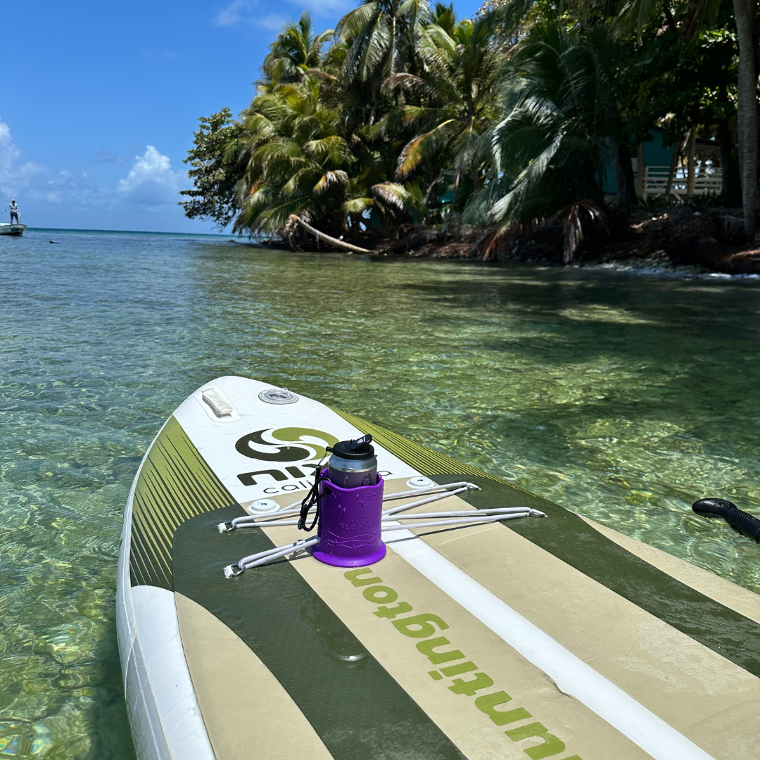 A green and beige paddleboard with a gray can in a purple cup holder on top of it in clear water with palm trees in the background.