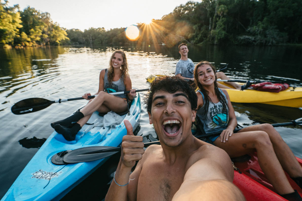 Four young adults enjoy a sunny day on a calm lake, paddling in colorful kayaks. The group includes a man in the foreground taking a selfie with a thumbs-up, a woman in a blue kayak, a woman in a red kayak, and a man in a yellow kayak.