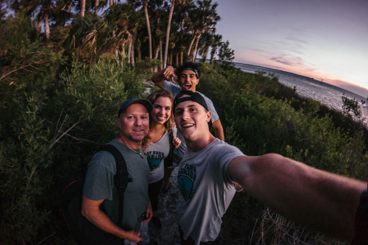 Four people taking a selfie outdoors on a cliffside path at sunset, with green foliage, palm trees, and the ocean in the background.