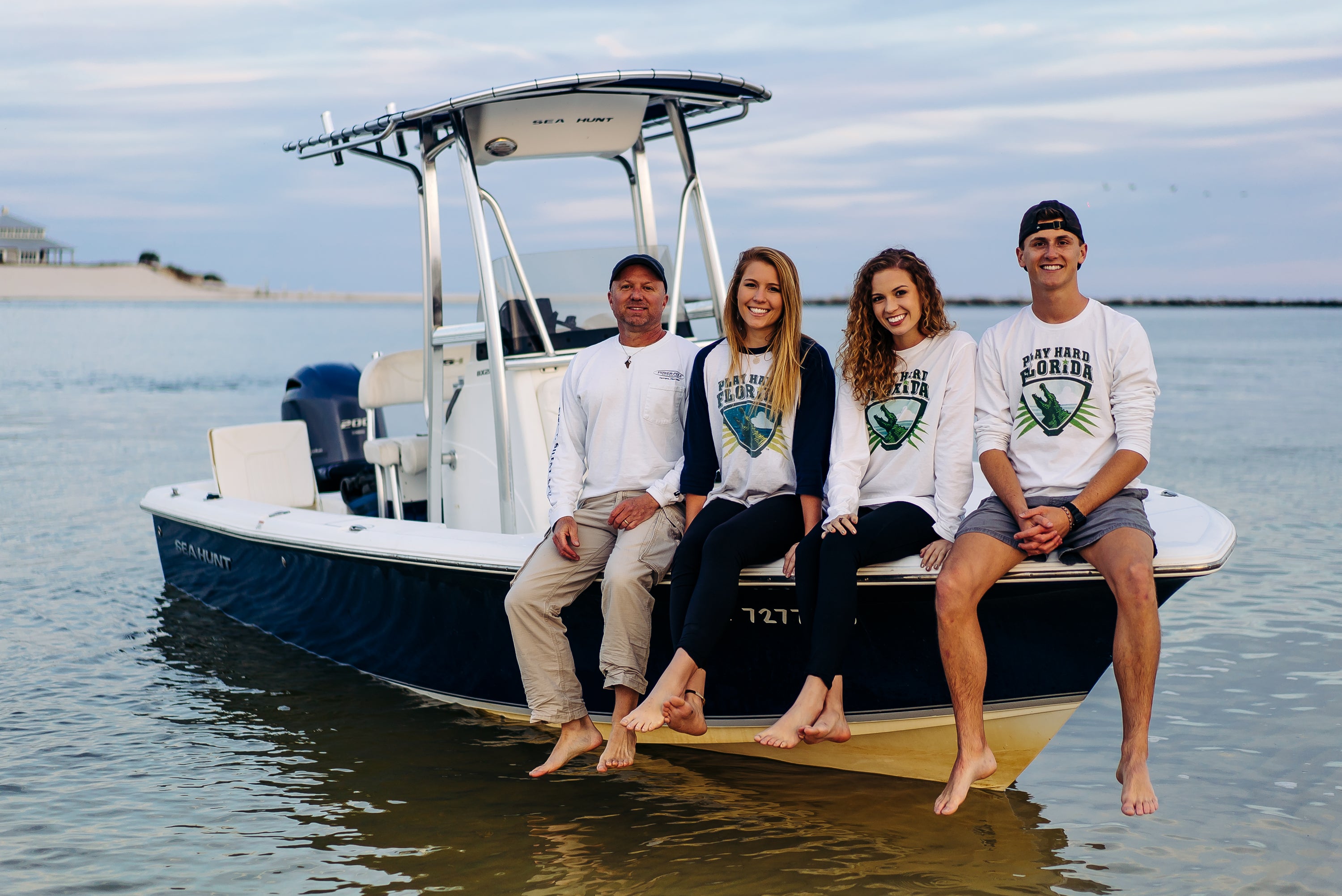 Four people sitting on the bow of a boat in the water, wearing matching white long-sleeve t-shirts with a green and blue 'Play Hard Florida' graphic on them.