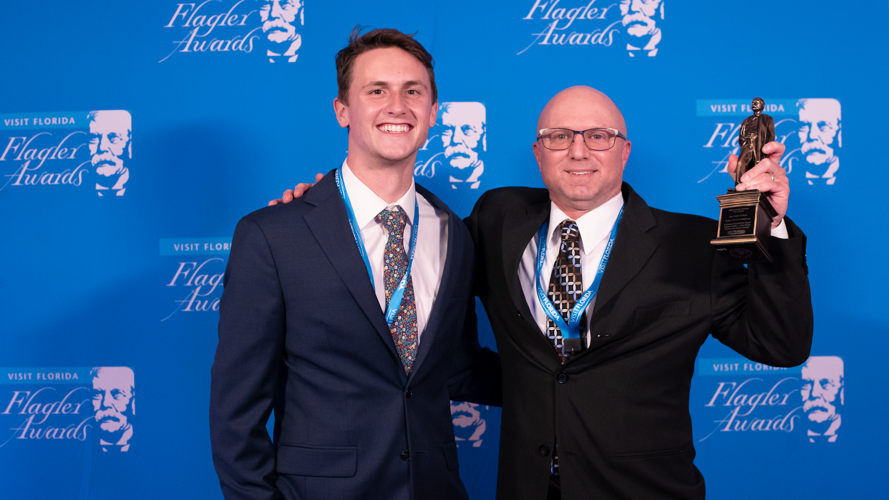 Two men in suits standing in front of a blue 'Visit Florida Flagler Awards' backdrop, with the man on the right holding a trophy.