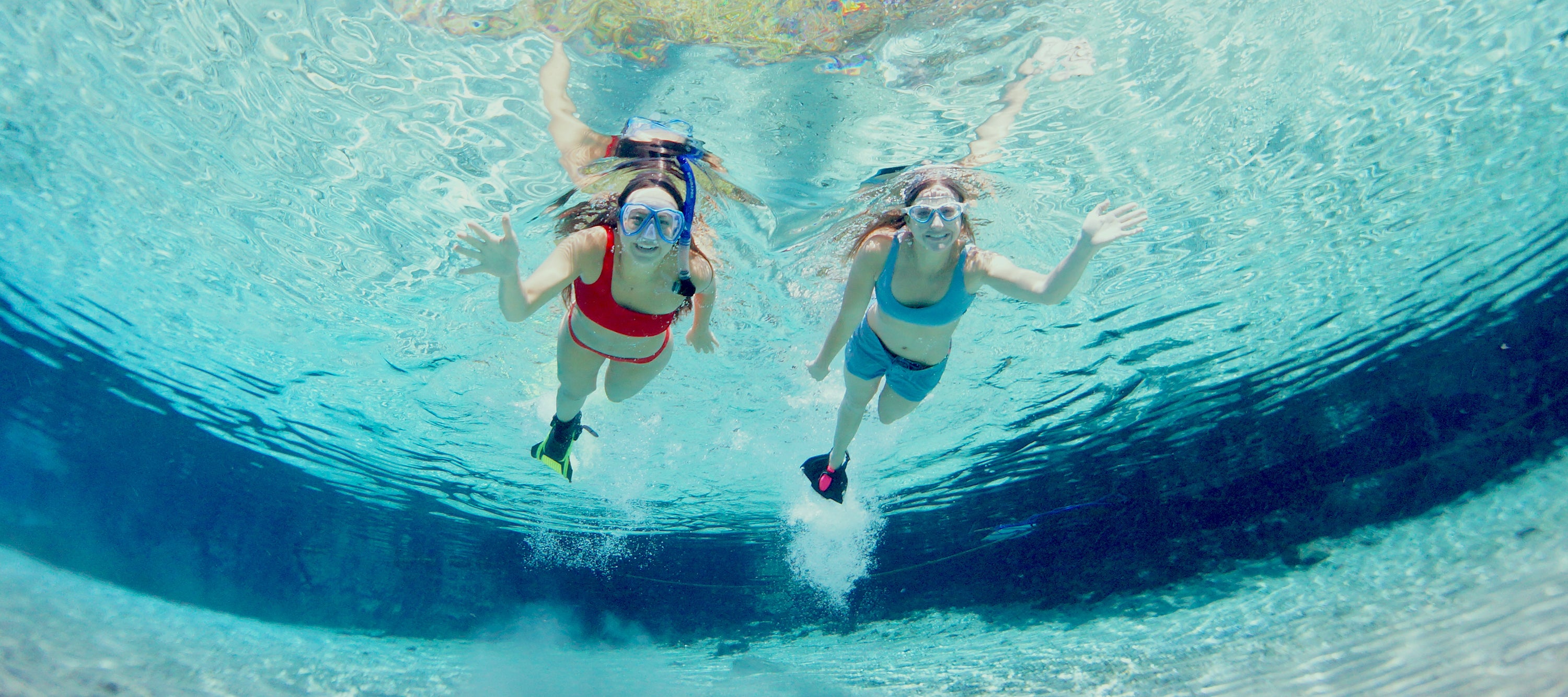 Two girls are snorkeling in light blue water while smiling and waving at the camera.