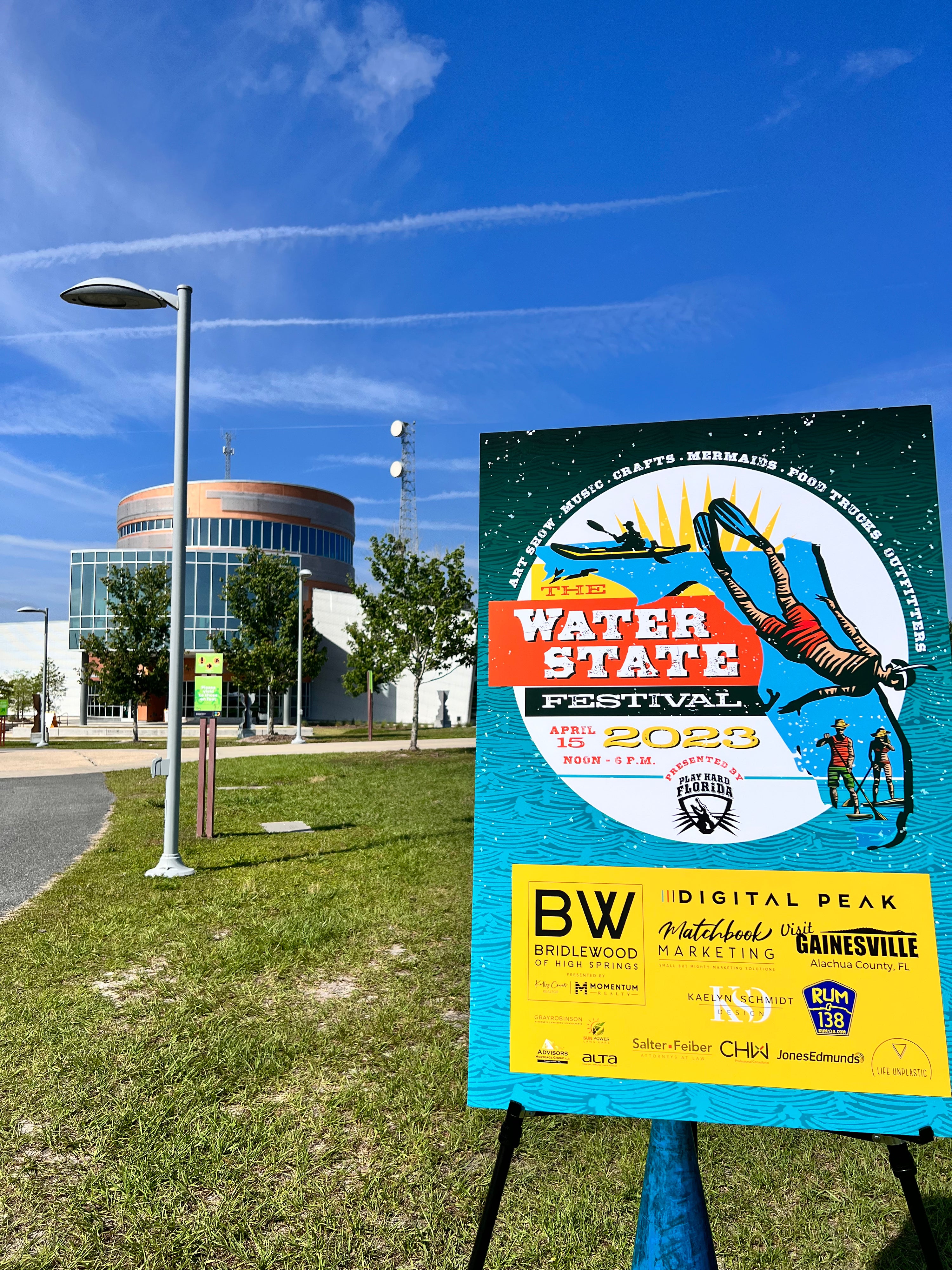 A promotional sign for The Water State Festival 2023 stands on a grassy lawn outdoors in front of the Cade Museum. The sign features a blue and yellow design with an illustration of a snorkeler on top of a blue Florida silhouette and sponsor logos.