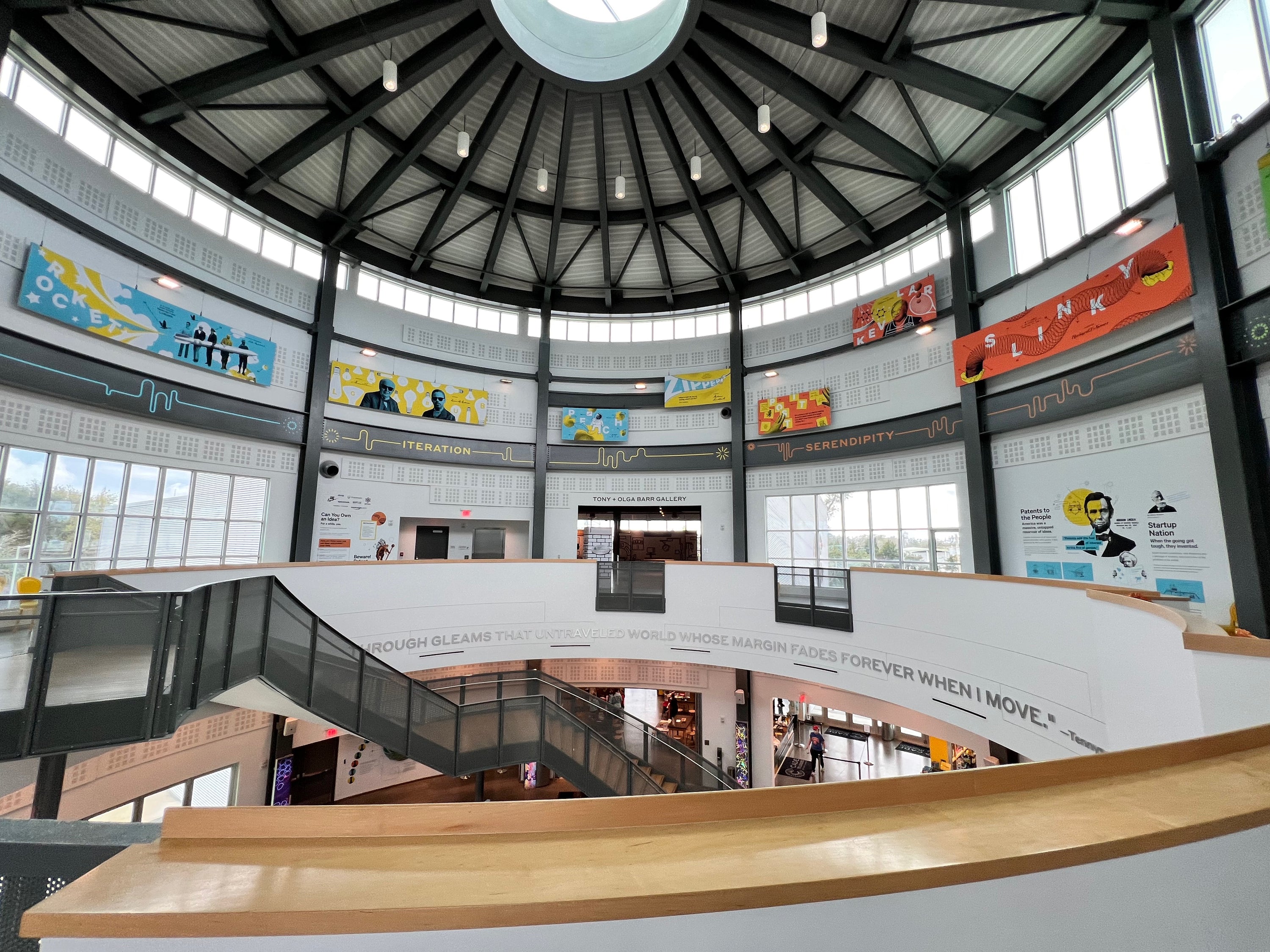 A high-angle view inside the Cade Museum, showing a large modern domed building with a circular atrium, a staircase, tall windows, and colorful wall murals around the upper levels.