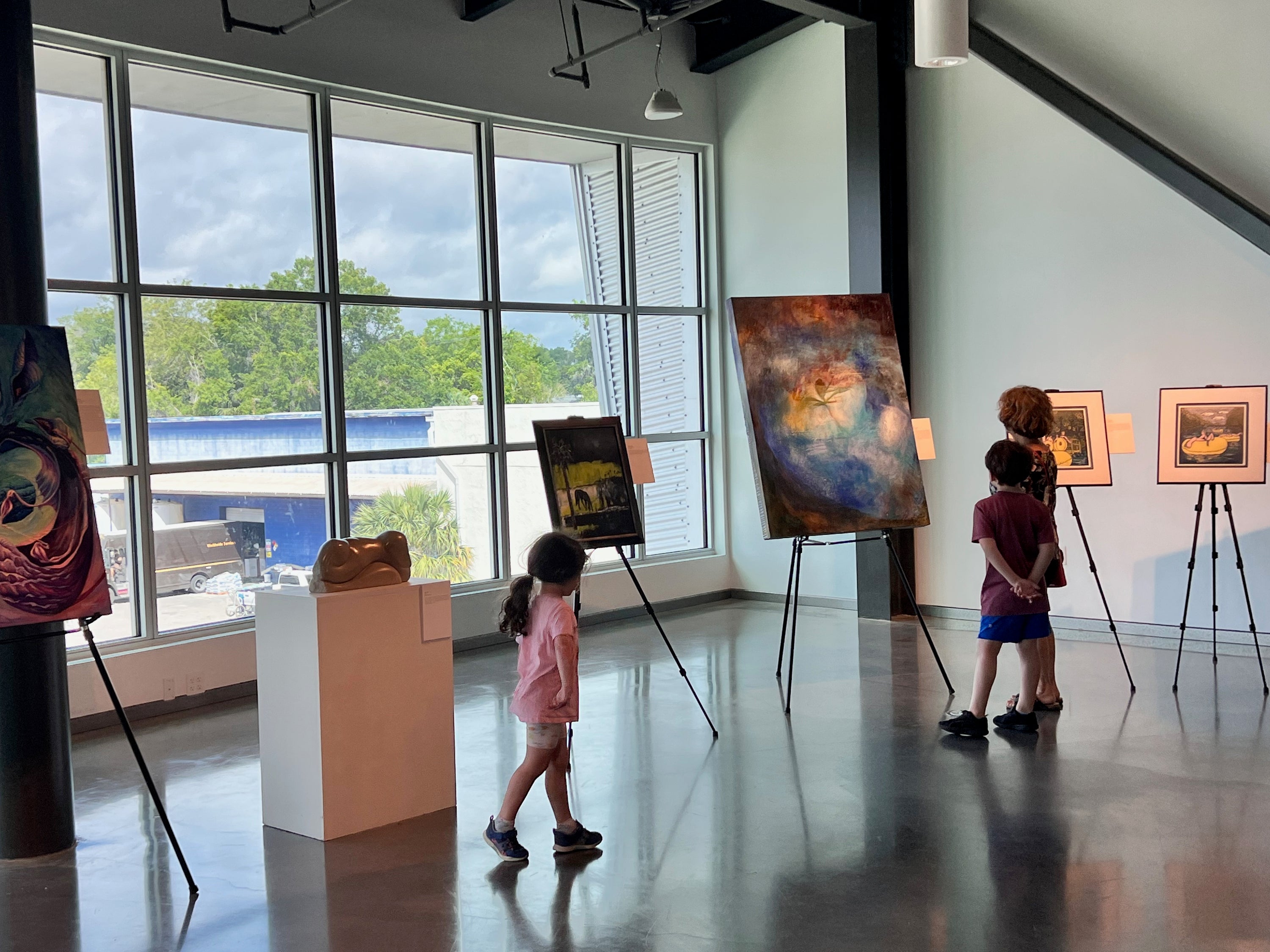 Children viewing a colorful abstract painting displayed on an easel in a modern art gallery space with large windows and other art pieces on display.