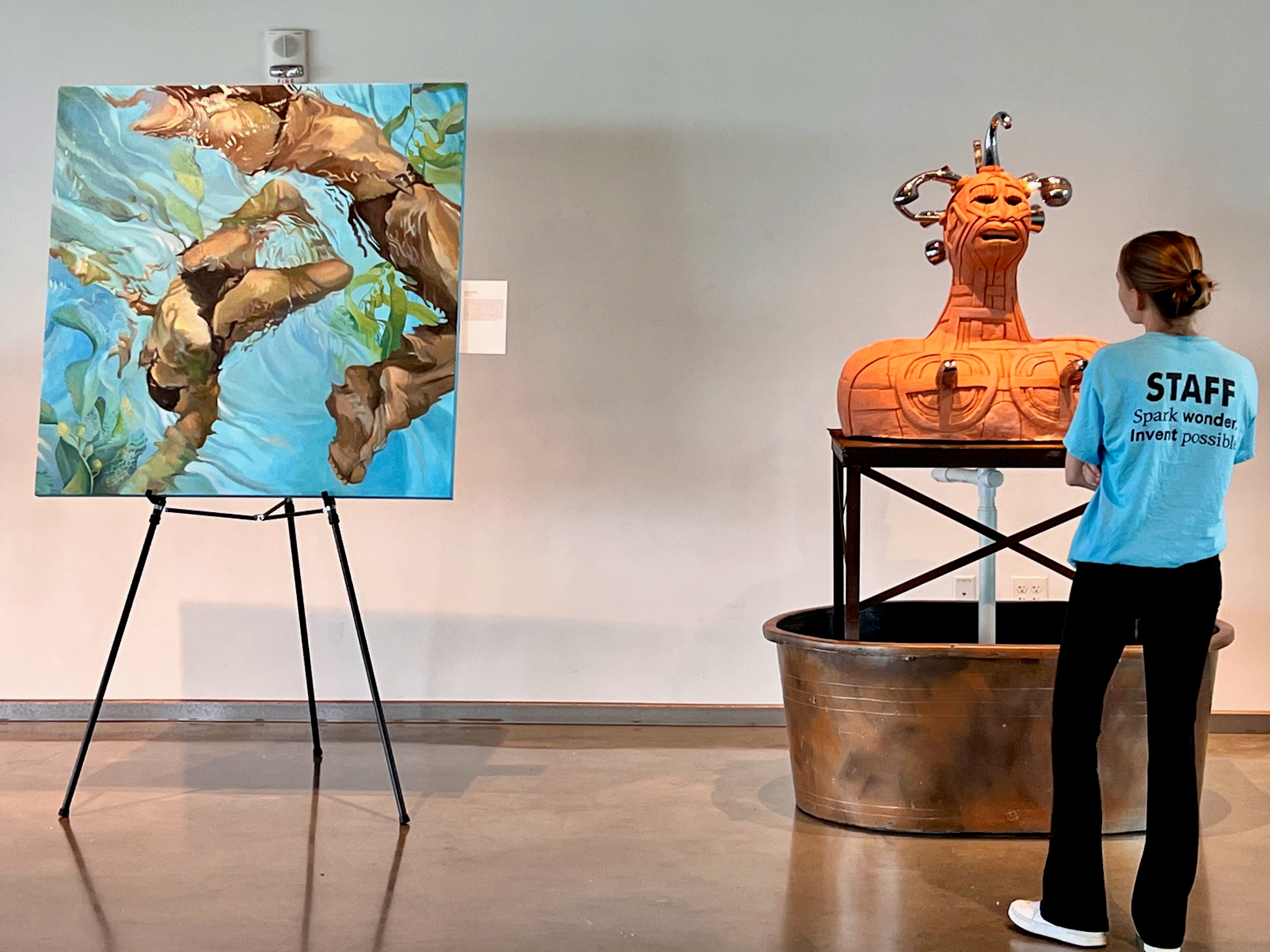 An art gallery interior shows two artworks: a colorful painting on an easel depicting women swimming in blue water, and a large orange carved bust sculpture on a pedestal in a metal tub. A person wearing a bright blue shirt with 'STAFF' printed on the back stands in front of the sculpture.