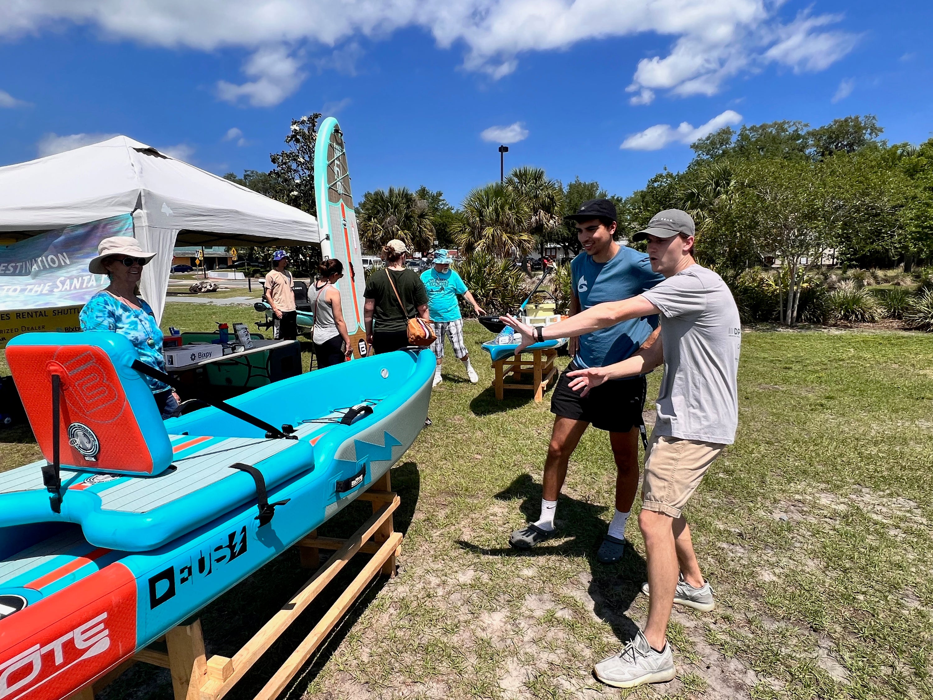 A bright turquoise and red sit-on-top kayak is displayed outdoors on a wooden stand at a sunny event, with several people standing nearby under a white canopy and trees in the background. A man is showing off the kayak to another man in the foreground.
