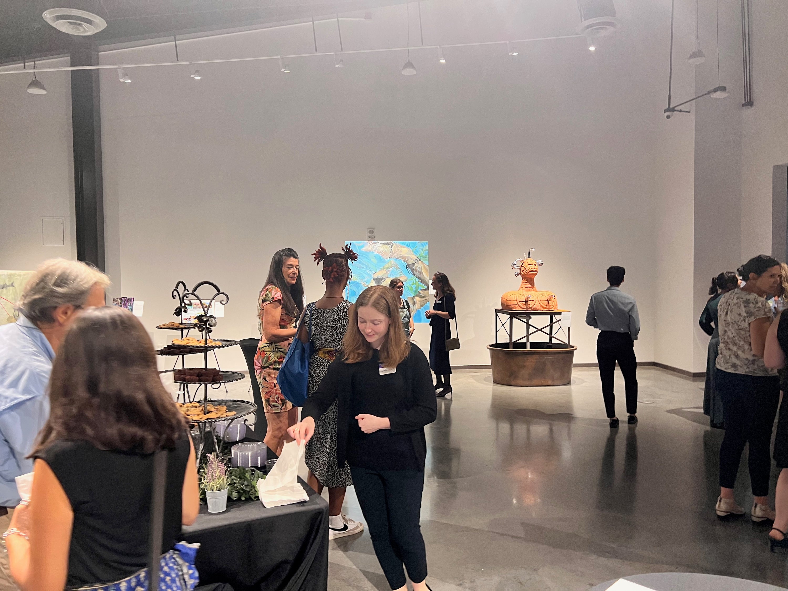 A group of people gathered in a gallery space with white walls and a polished floor, viewing art pieces including a colorful water painting and a large orange bust sculpture on a pedestal.