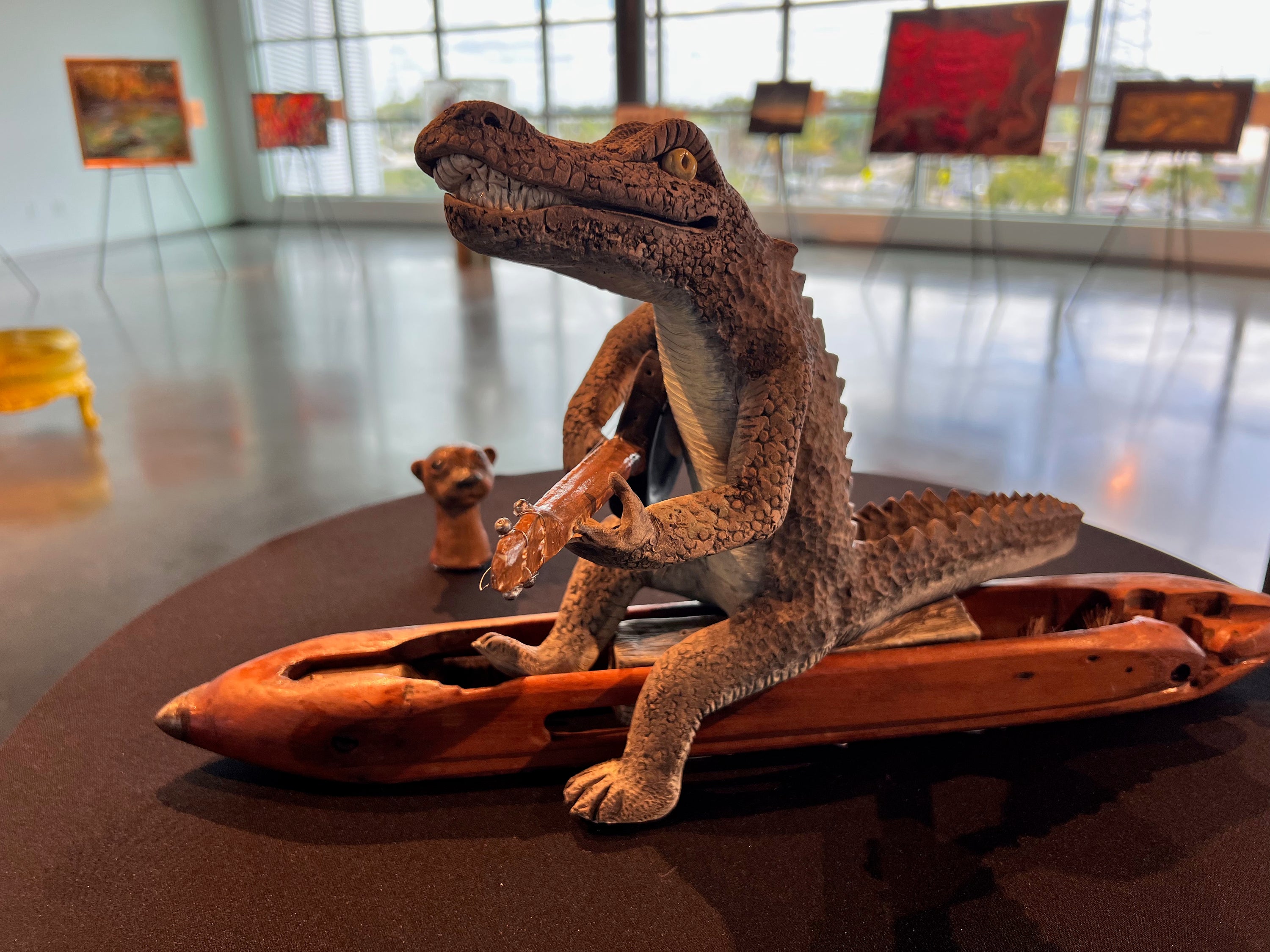 A detailed alligator figurine sitting in a carved wooden boat holding a guitar, displayed on a dark round table in a gallery or museum setting with art pieces displayed in the background.