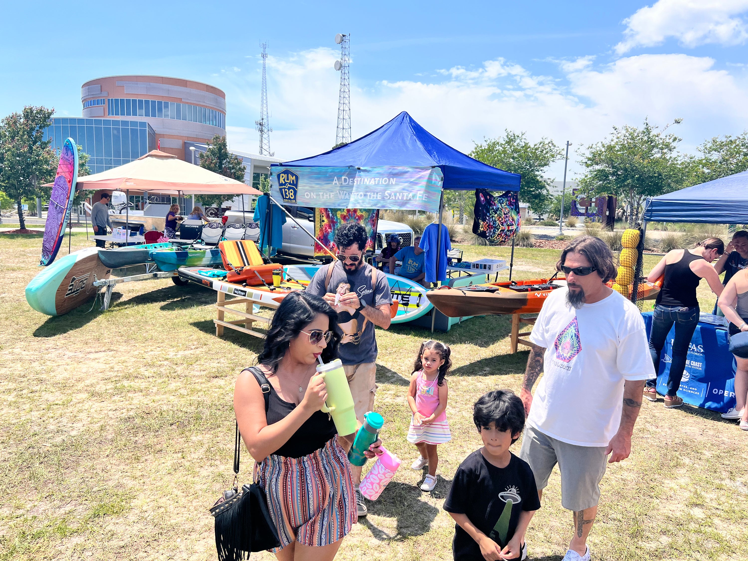 Outdoor event scene on a grassy field with several people gathered near canoes and paddleboards under blue and tan canopy tents. A woman in the foreground holds a green bottle, with a man in a white shirt and a boy beside her.