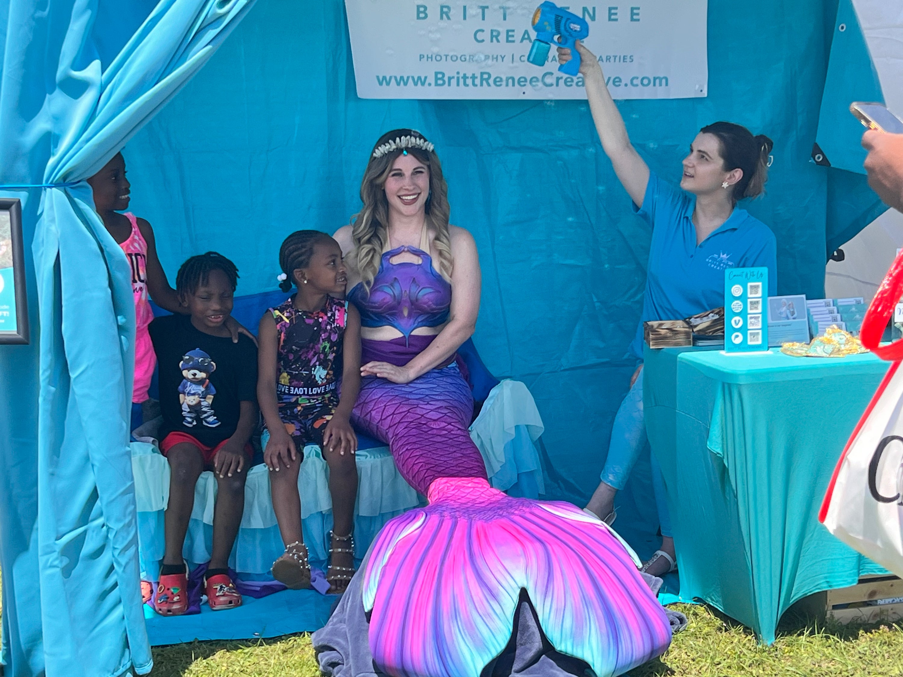 A woman in a purple and pink mermaid tail costume sits on a blue bench under a light blue canopy tent, sitting next to children, while another woman in a blue shirt blows bubbles with a bubble gun above them. A sign for Britt Renee Creative is visible in the background.
