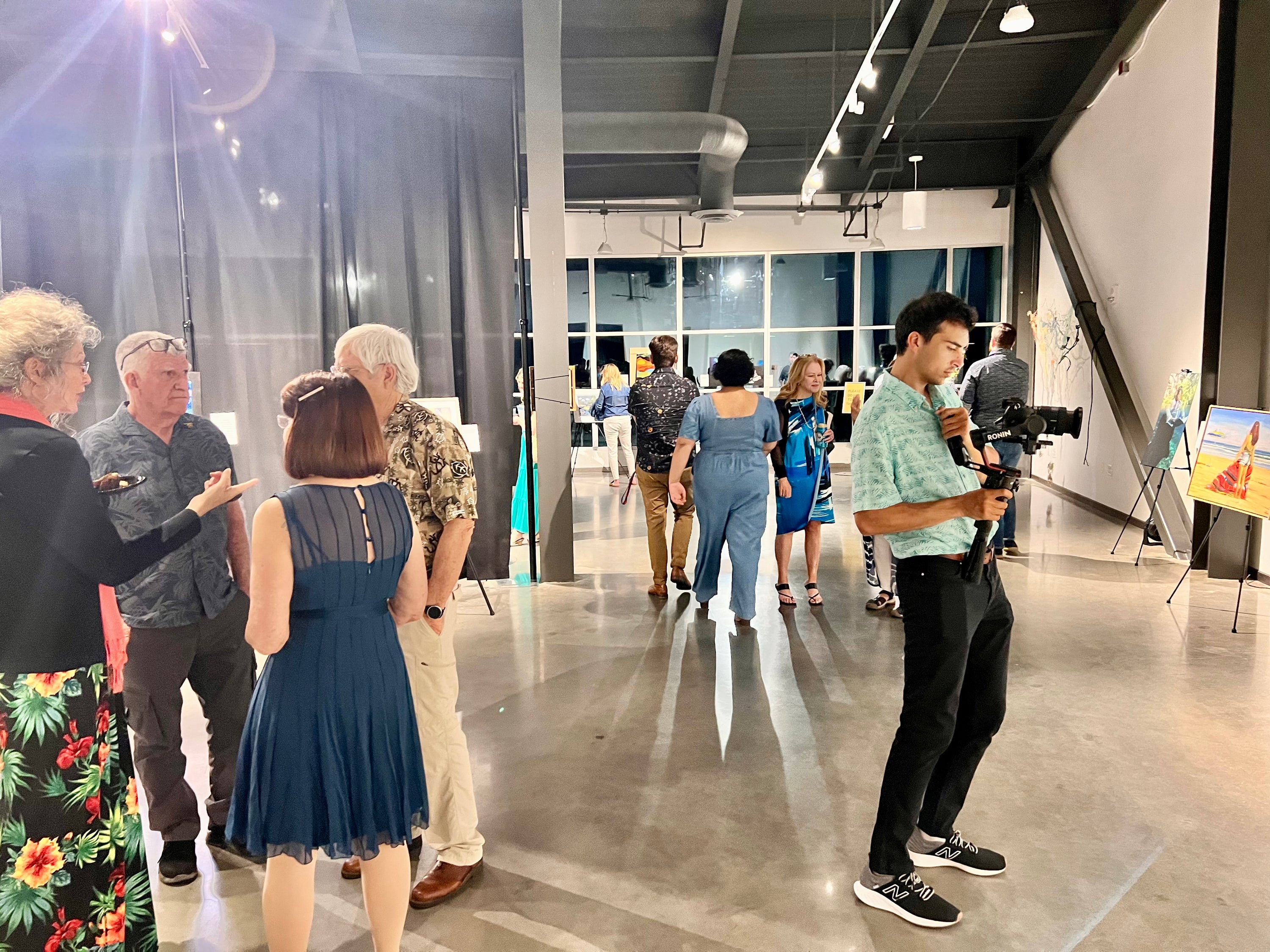 A group of people gathered in a modern art gallery with high ceilings, windows, and art displays. A man on the right holds a camera, while others converse and view artwork in the background.