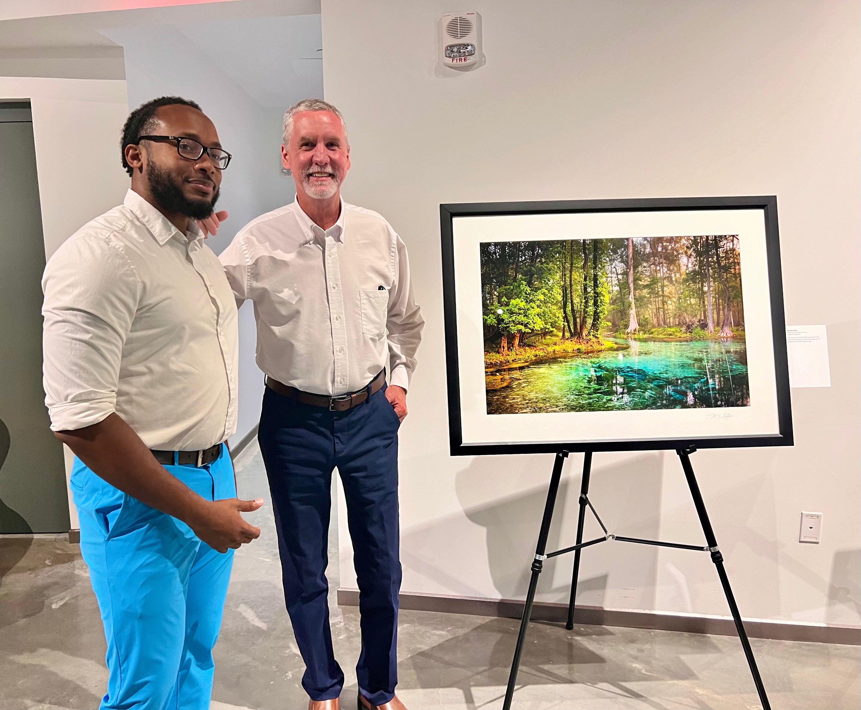 Two men standing indoors beside a framed landscape art print on a tripod easel. The artwork shows a forest with a turquoise spring.