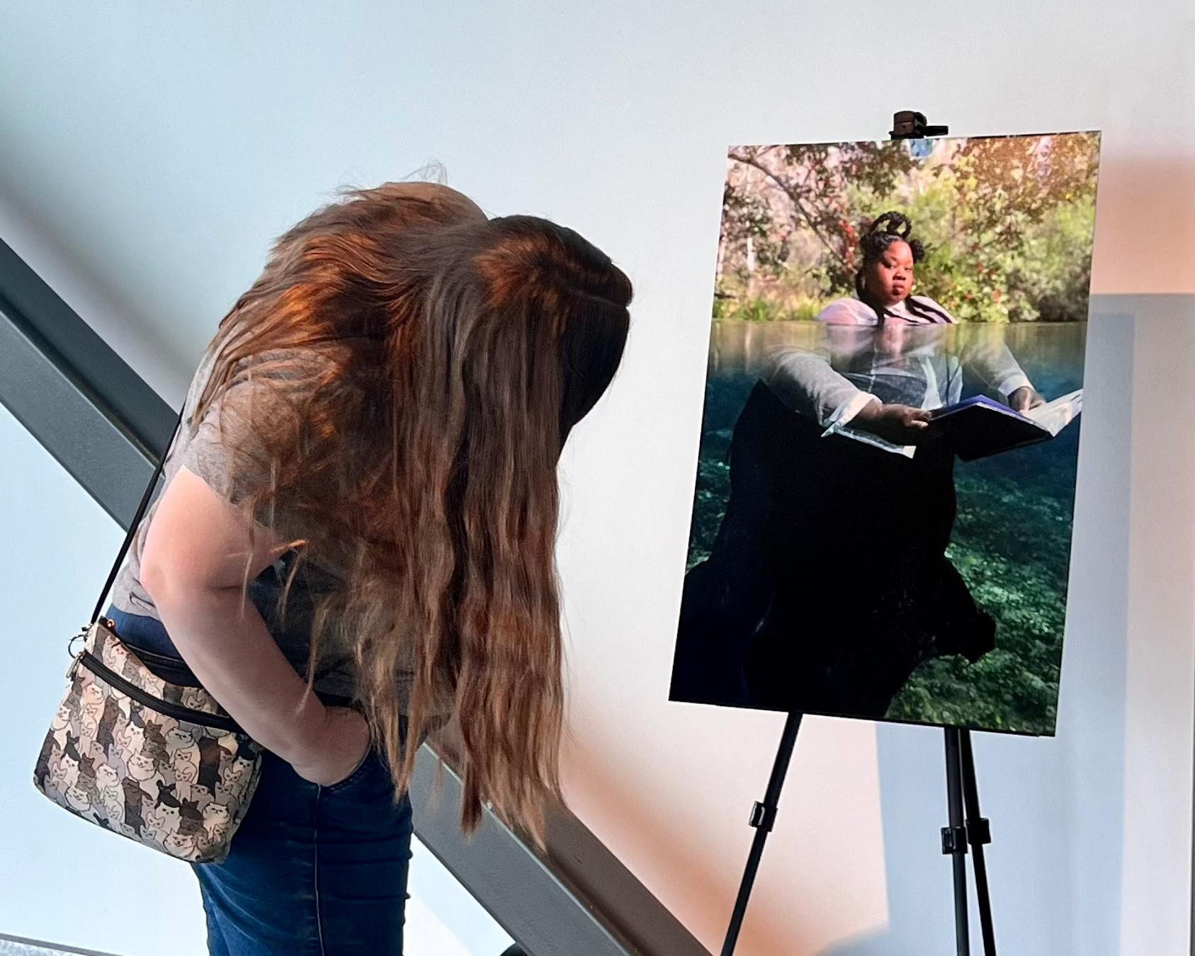 A woman with long brown hair looks at an easel displaying a photographed scene of a woman in a dress reading in a body of water with the book underwater. The woman is wearing a gray patterned top, dark blue jeans, and a patterned shoulder bag.