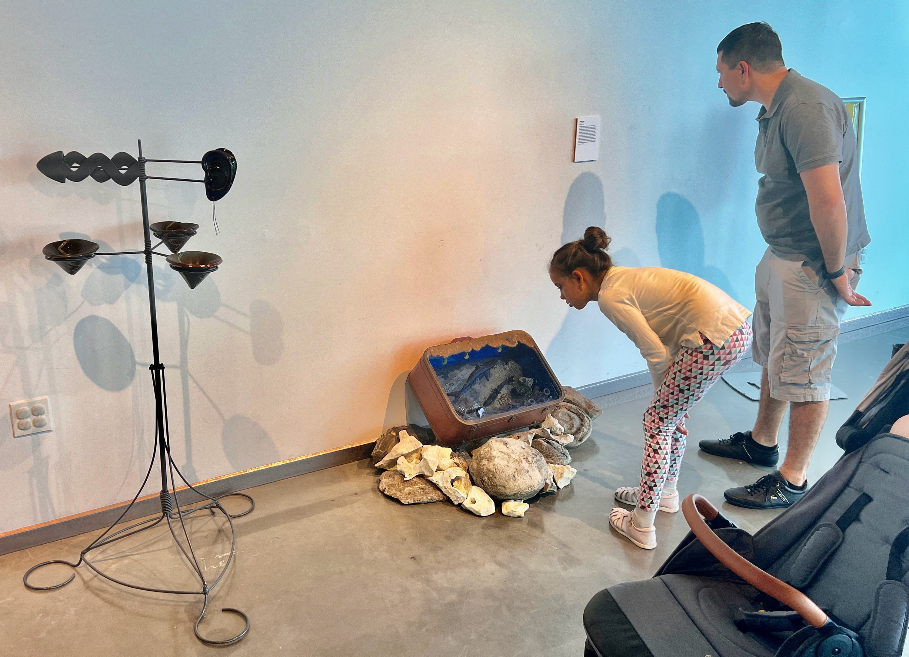 A young girl and a man look at an art piece in a modern art gallery with white walls. The art piece is a sculpture of a briefcase with sharks in water showing through the front, surrounded by shells and rocks. The girl is in white attire and crouching, while the man is in a gray polo shirt and khaki shorts.