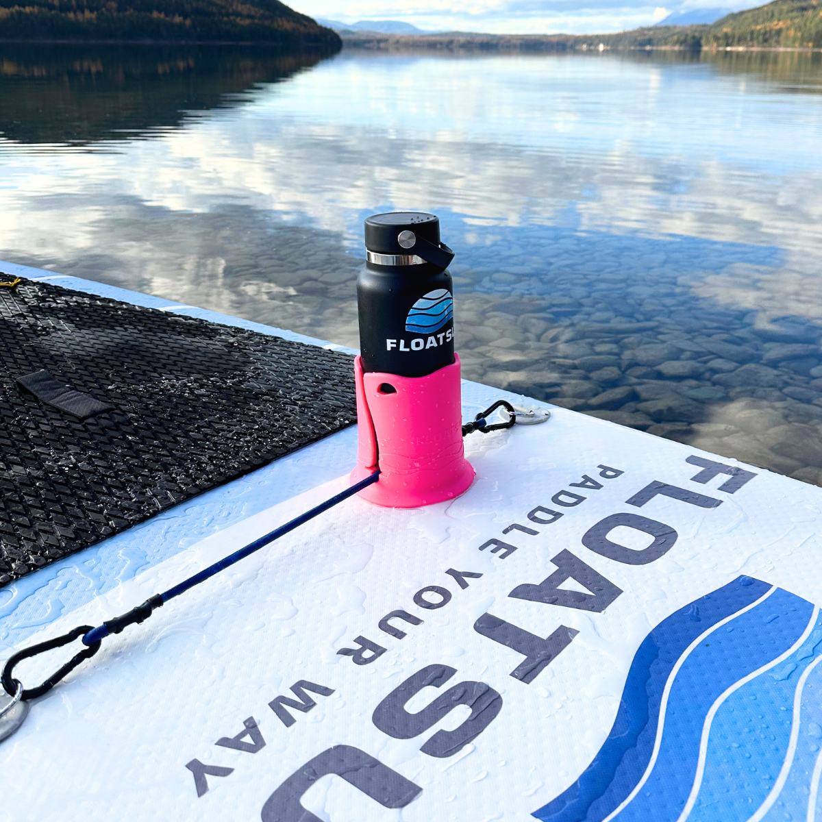A black water bottle with the Floatsup logo on it in a pink cup holder on a white paddleboard in the water of a lake with mountains in the background.