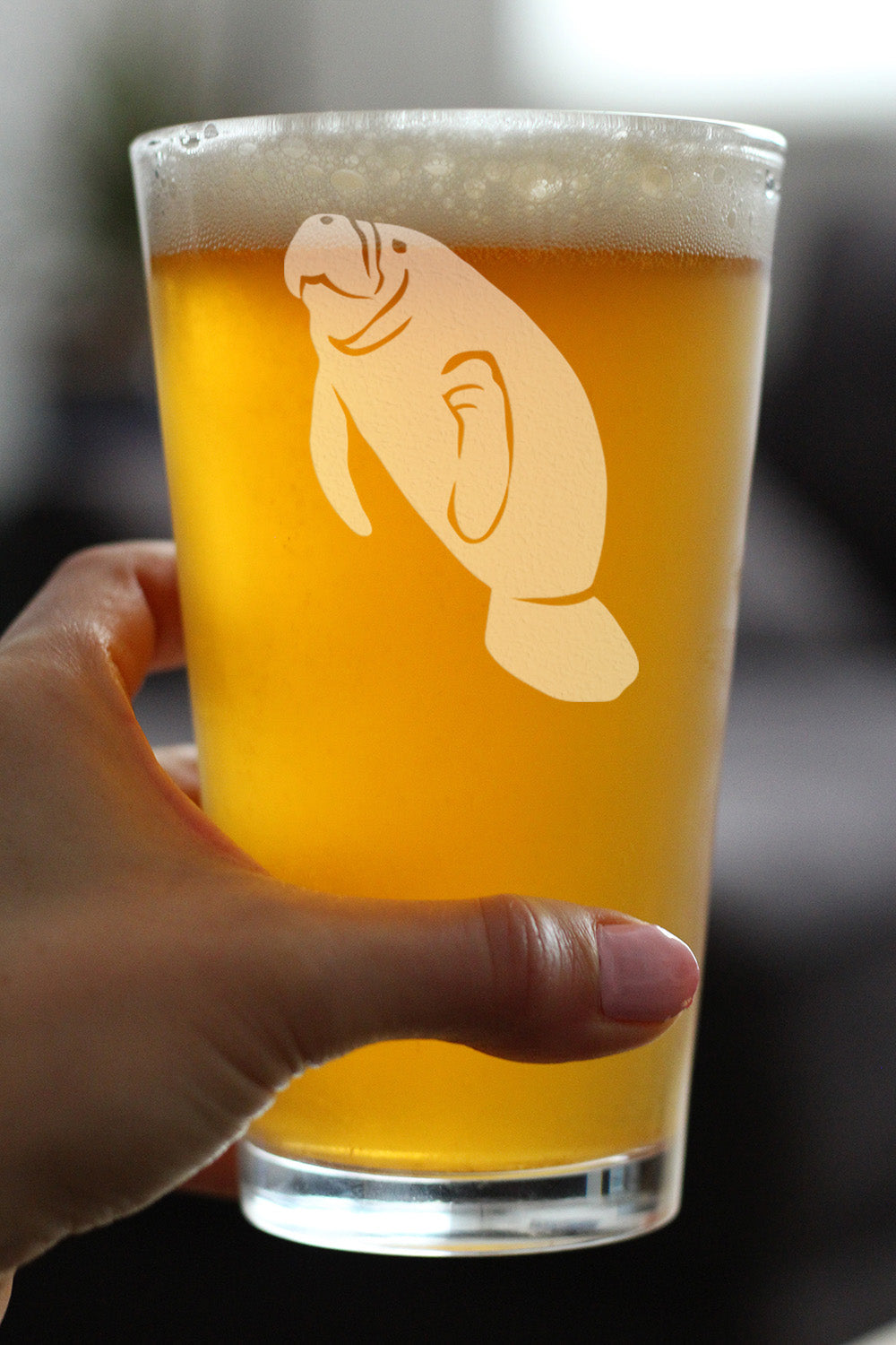 A hand holding a clear glass with an etched manatee design on it that is full of beer on a blurred background.