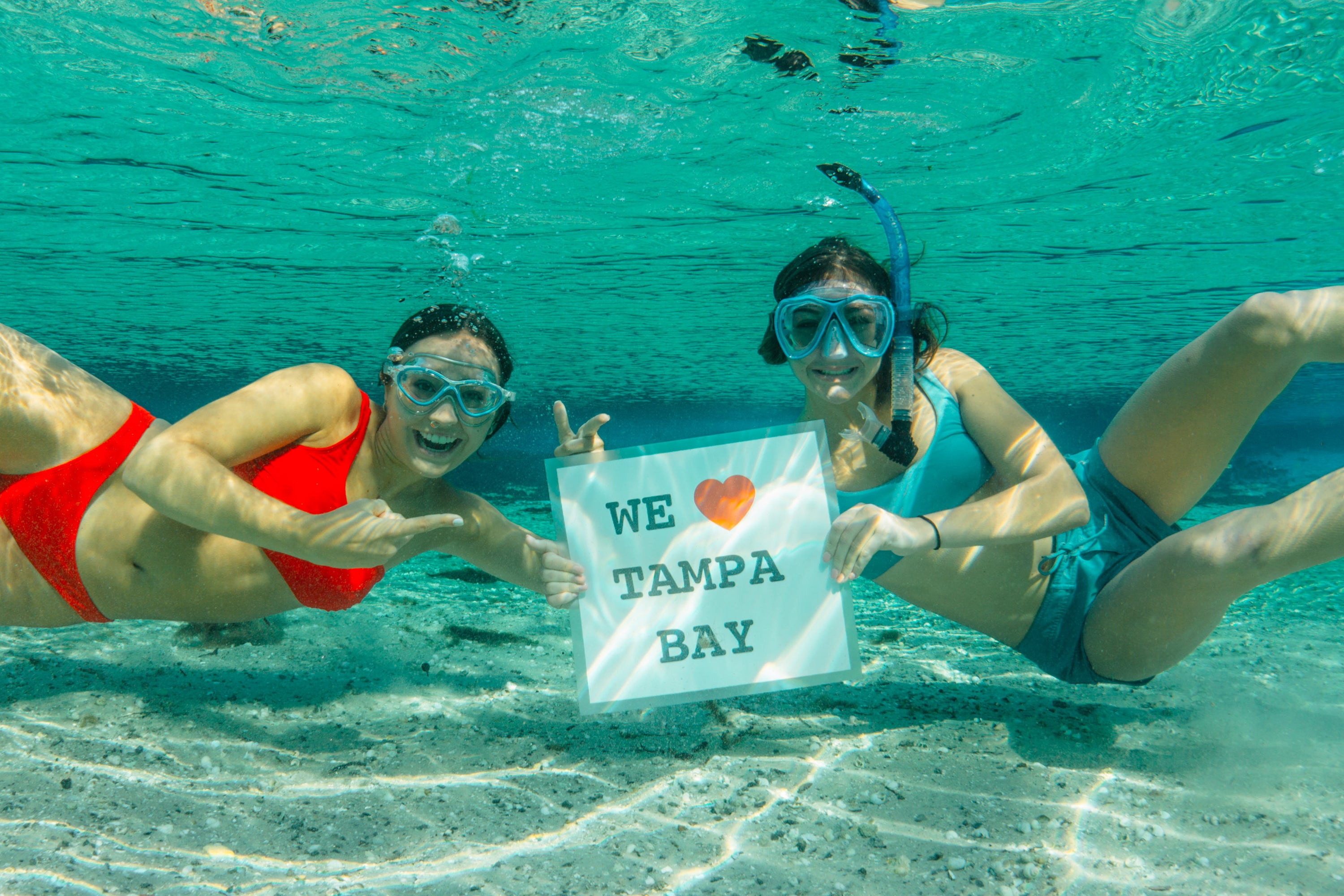Two women snorkeling underwater in clear turquoise water, each wearing a mask and holding up a white sign that reads 'WE ♥ TAMPA BAY'.