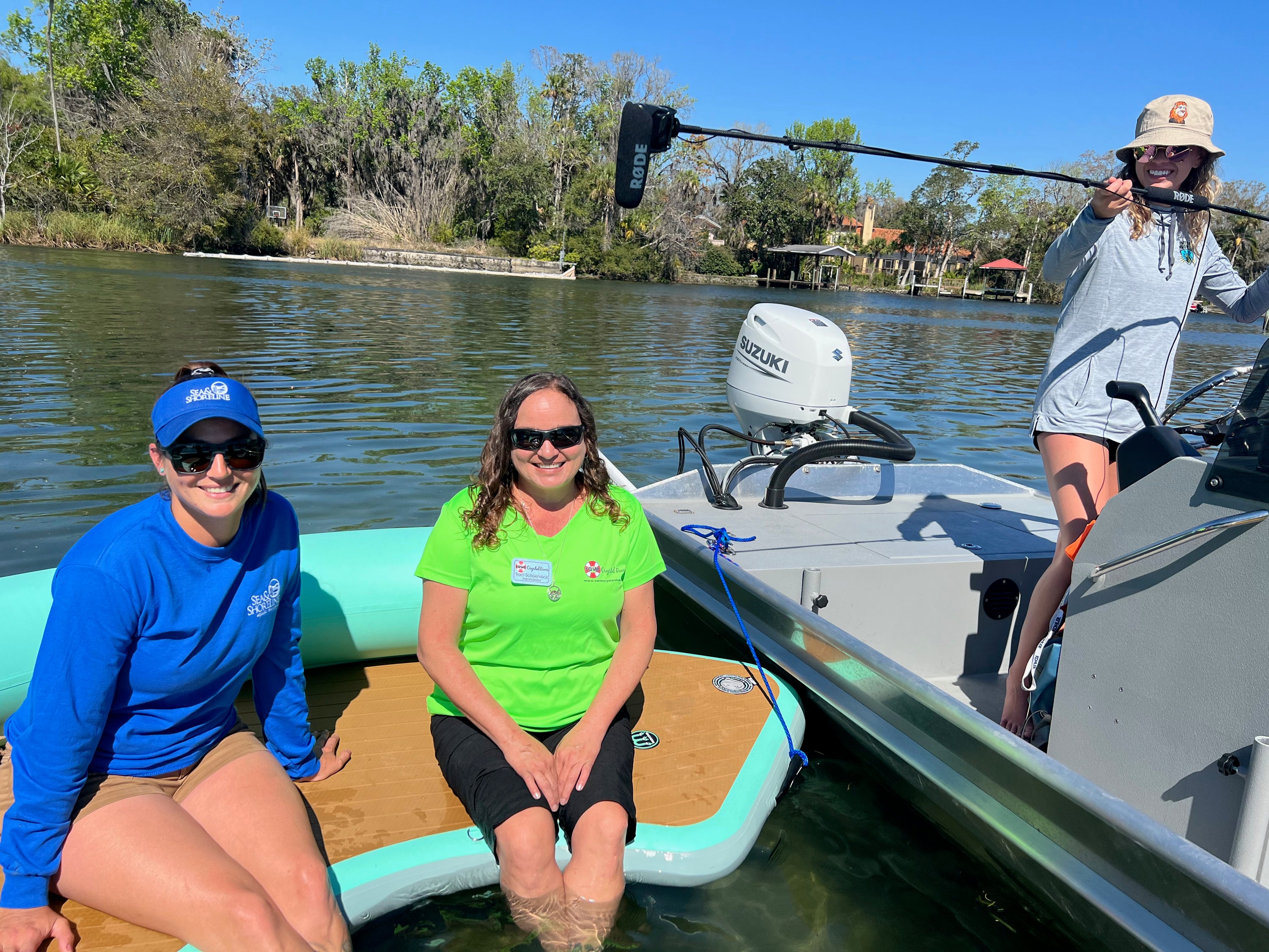 Three women on a boat on a river, with two women sitting on a turquoise and tan inflatable pad in the water, while the third woman is on the boat holding a microphone. A Suzuki outboard motor is visible in the background, along with trees and water.